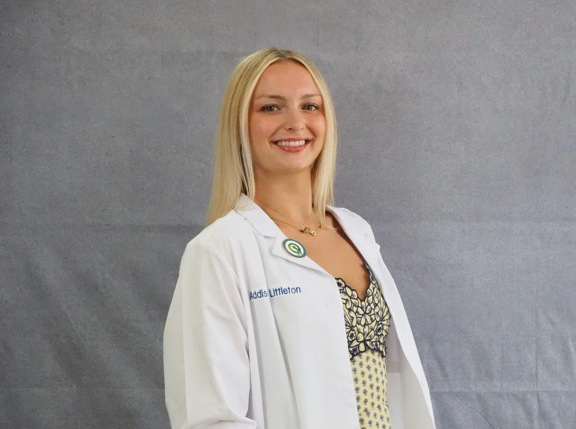 Smiling woman in a white lab coat with a logo, standing against a gray background.
