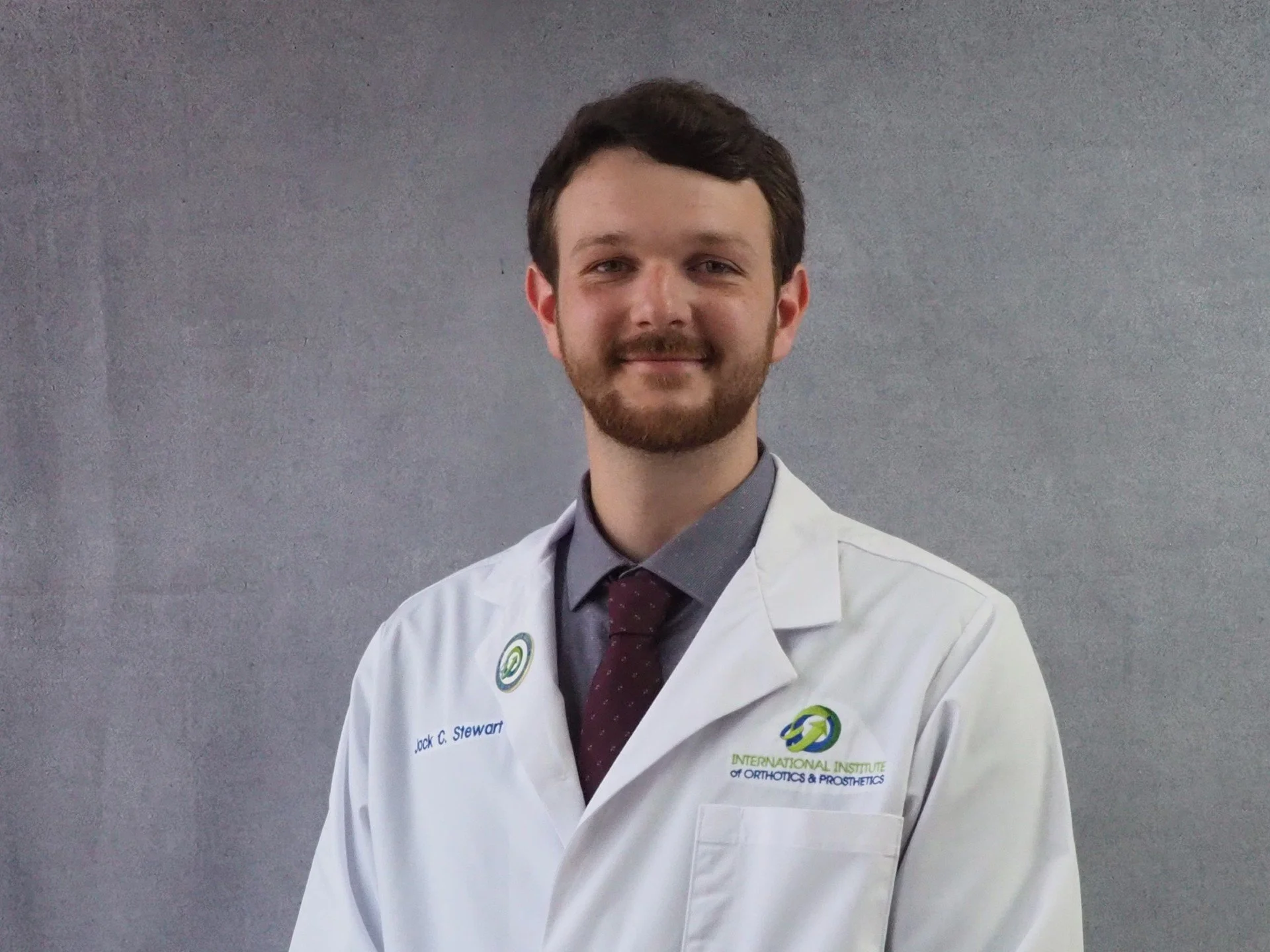 Man in a white lab coat, smiling, with brown hair, standing against a gray background.