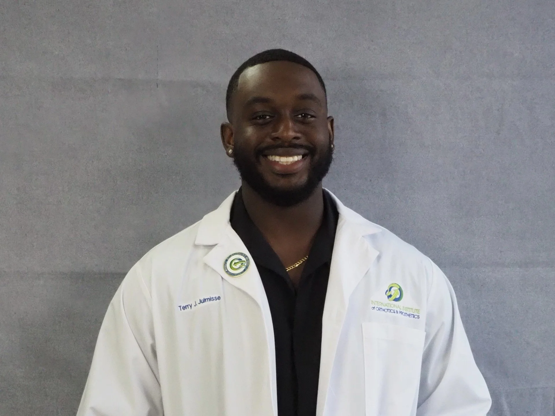 A person smiling, wearing a white coat with a logo and name tag, against a gray background.