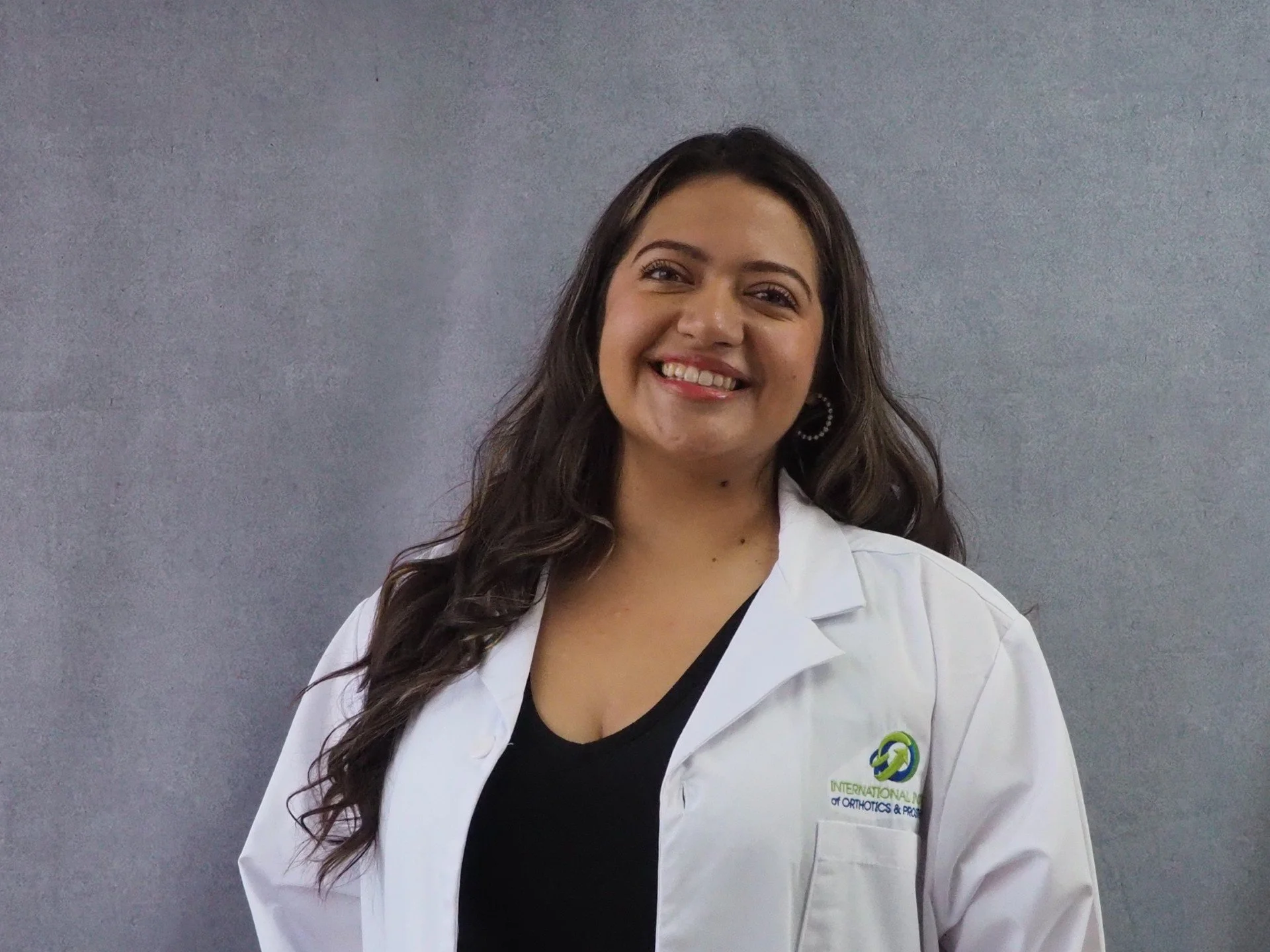 Person in a white lab coat with logos, smiling against a gray background.