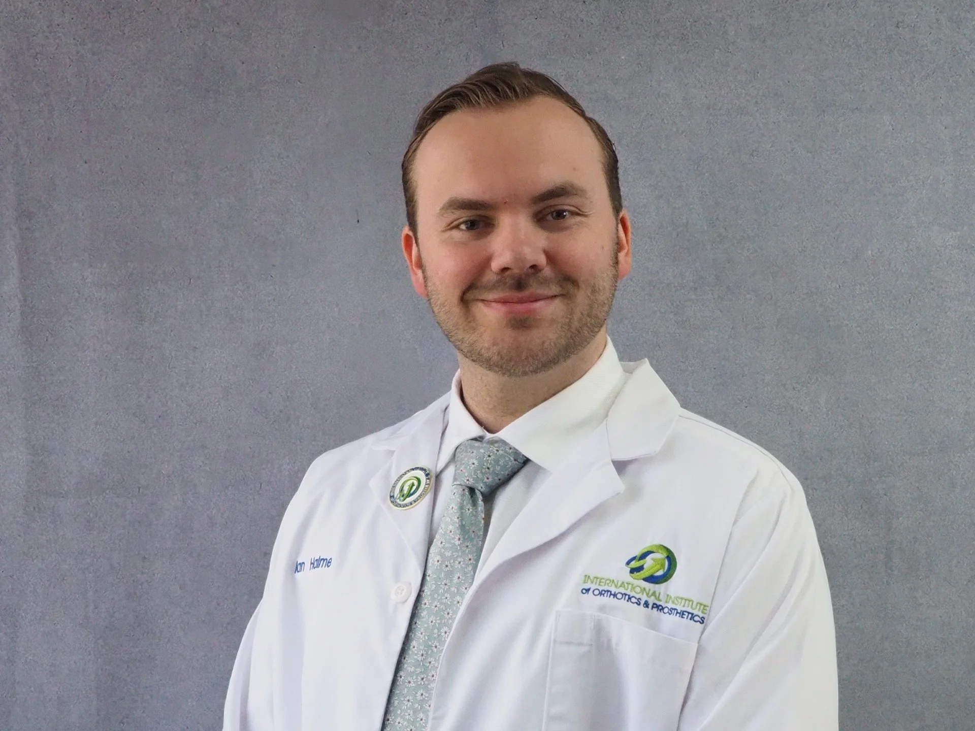 Smiling person in a white lab coat with a logo, standing against a neutral background.