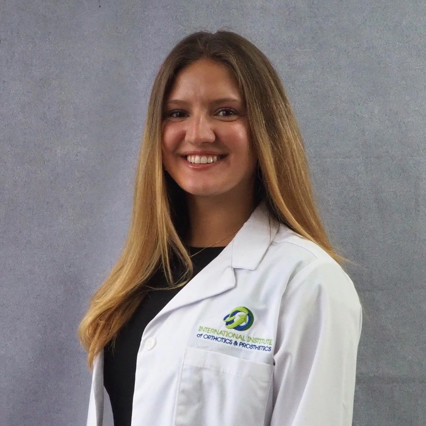 A person smiling, wearing a white coat with a logo and name tag, against a gray background.
