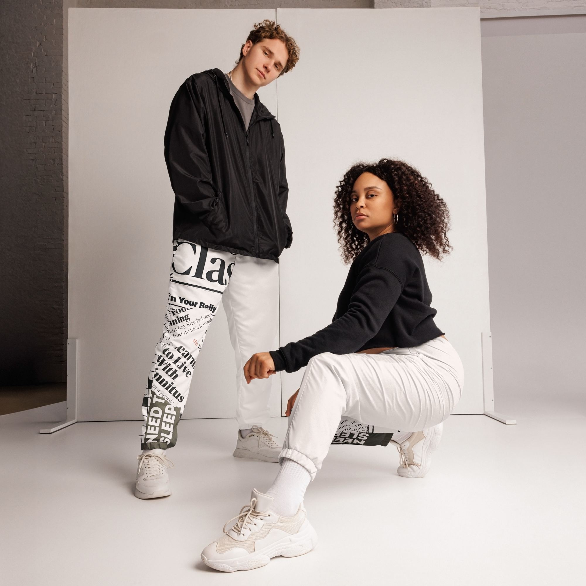 Two young adults, one male and one female, posing in front of a white background with a black brick wall on the left. The male has curly hair, wears a black jacket and white pants with black text, and stands with hands in his pockets. The female has curly hair, wears a black sweatshirt and white pants, and is squatting with one knee on the ground, looking at the camera.