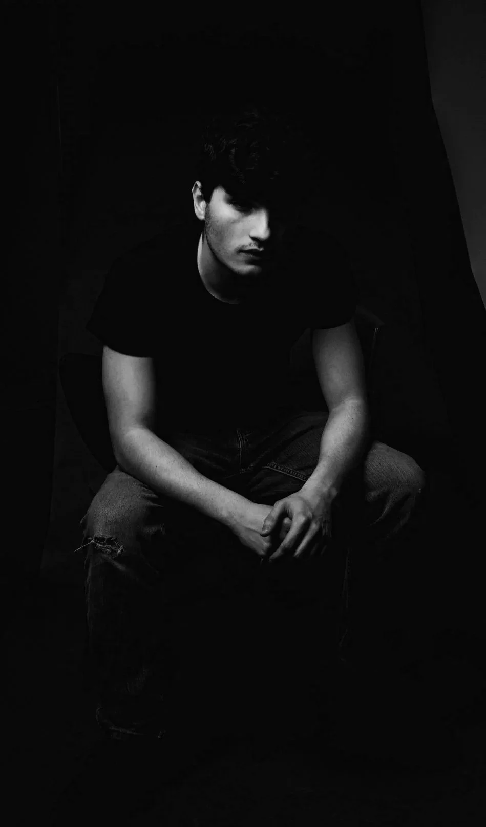 A young man with dark curly hair, wearing a black t-shirt and ripped jeans, sits in a dark room with shadows, looking at the camera.