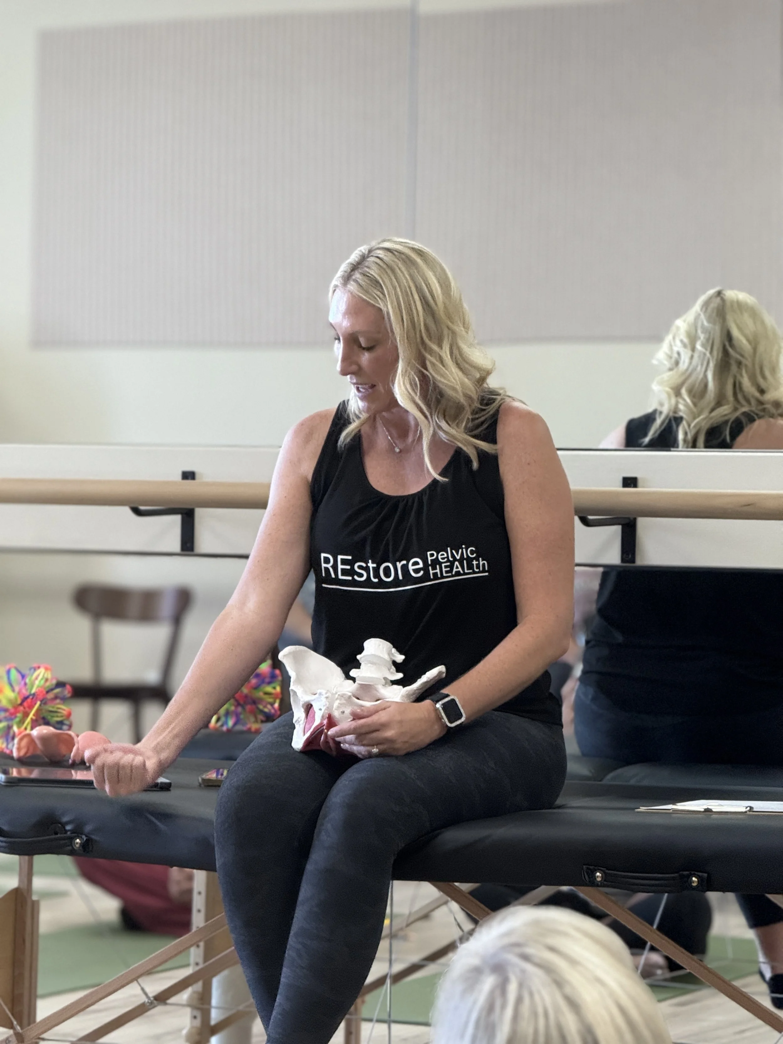 A woman with blonde hair, wearing a black sleeveless shirt that reads "REstore Pelvic HEALTH," sitting on a medical table, holding a pelvis model, in a classroom or workshop setting.