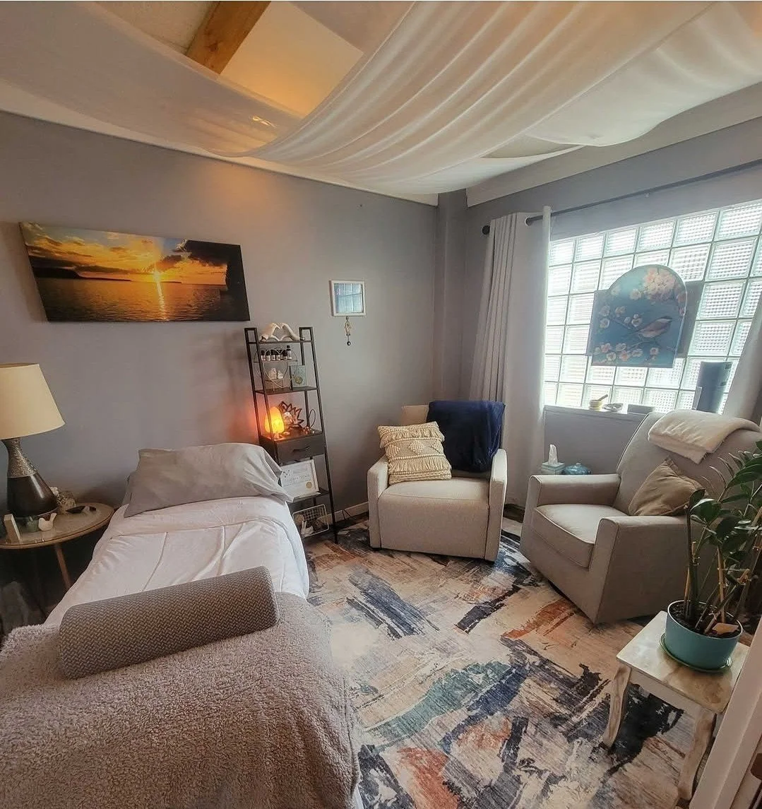 A cozy living room with a bed or daybed, an armchair, two plush chairs, a rug with warm colors, a window with curtains, and decorative items like a plant and artwork.