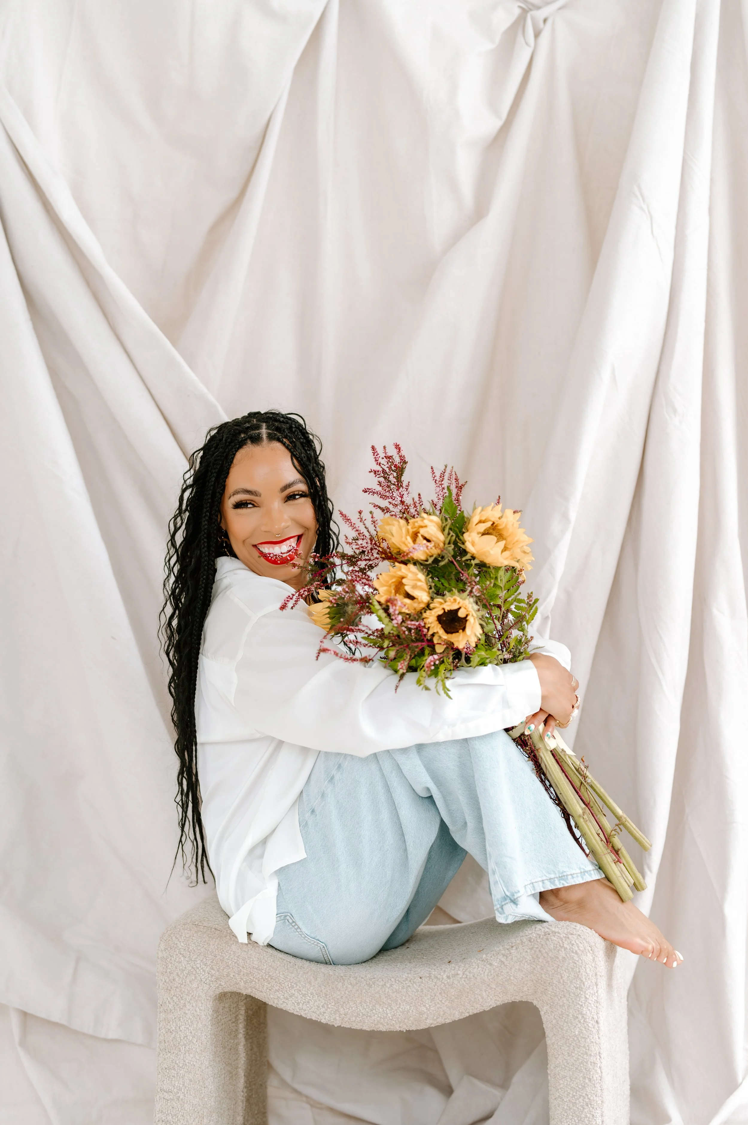 A woman with long black curly hair, wearing a white shirt and light blue jeans, is sitting on a beige stool, holding a bouquet of sunflowers and pink flowers, against a draped white fabric backdrop, smiling and looking at the camera.