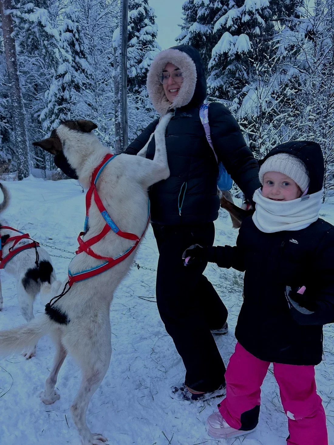 A woman and a young girl dressed in winter clothing in a snowy forest, with the woman holding a dog that is standing on its hind legs, partially leaning on her.