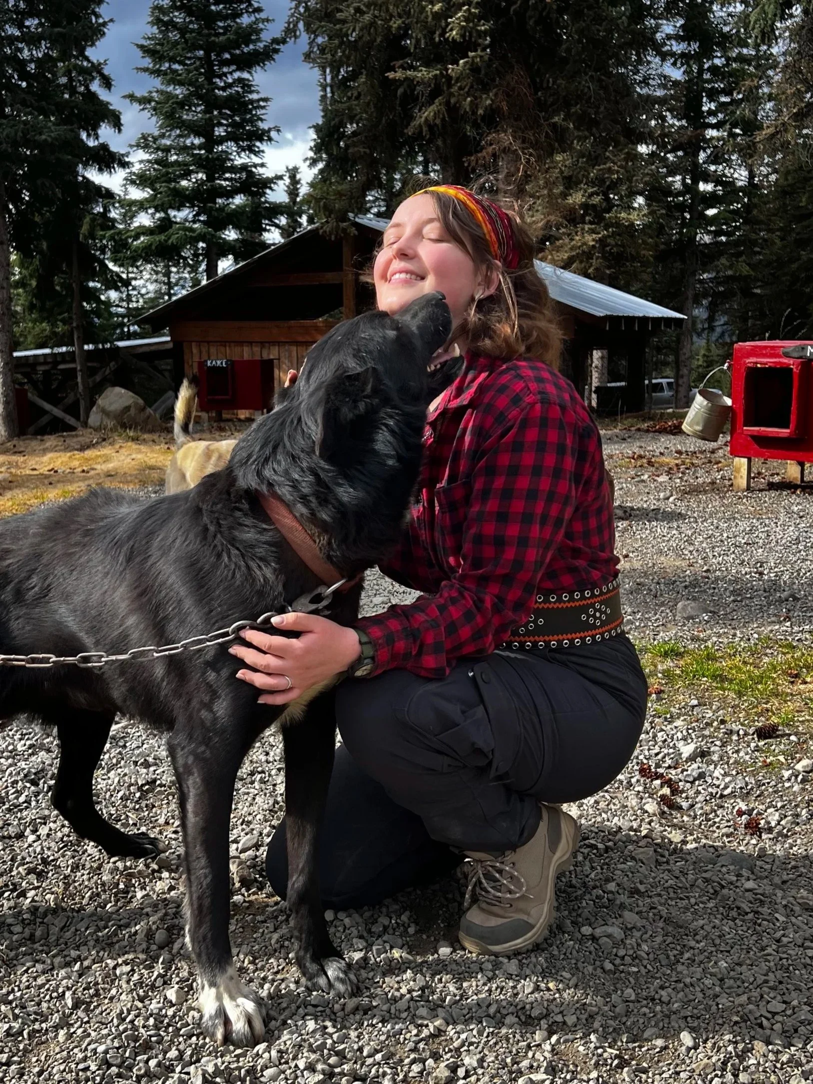 A woman kneeling and hugging a black and white dog outdoors with trees and a wooden building in the background.