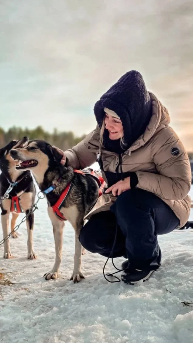 A woman in winter clothing kneels in the snow, smiling while petting two sled dogs wearing harnesses.