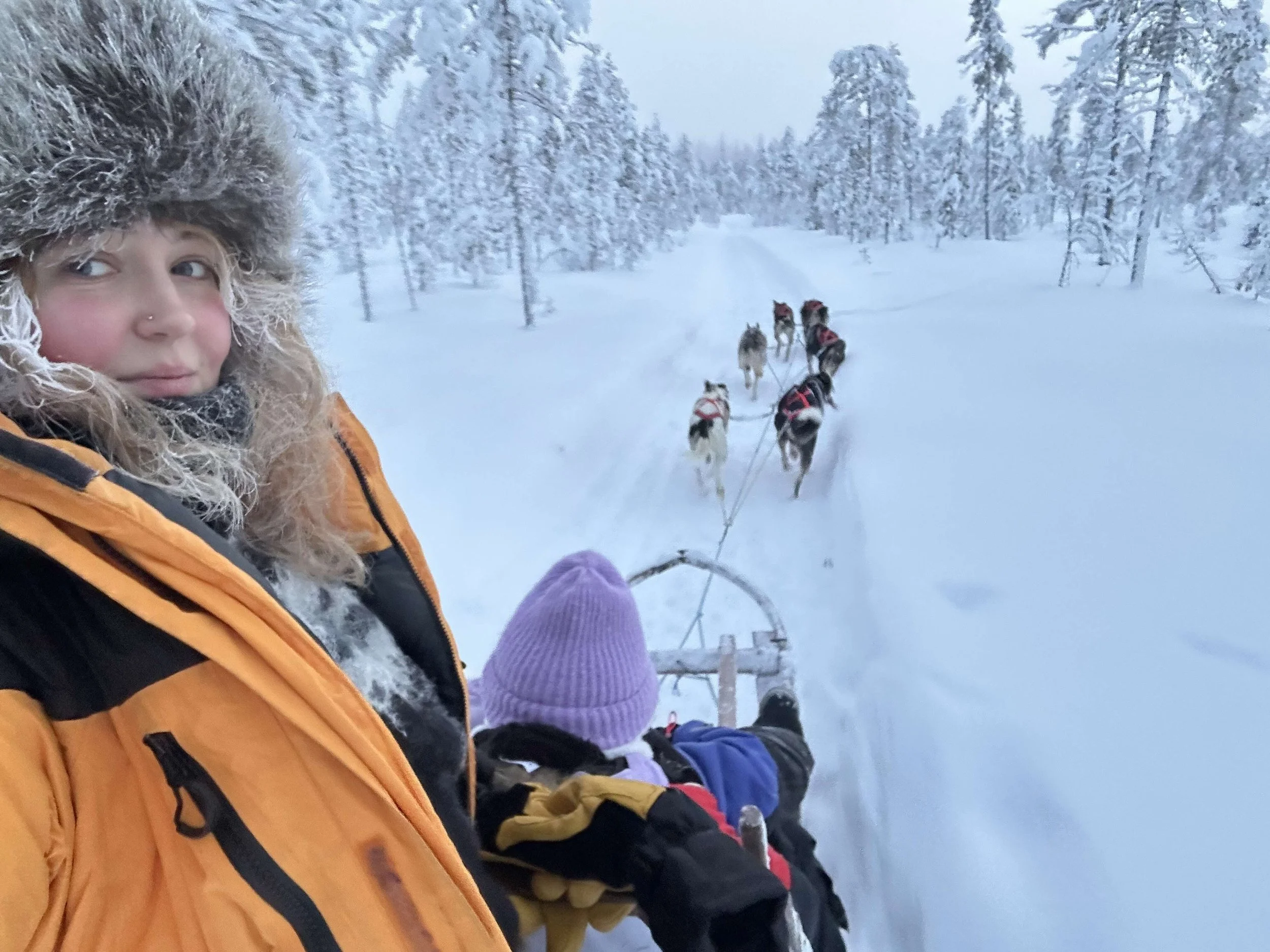 A smiling woman with wavy brown hair, wearing a green knit beanie and a dark jacket, is taking a selfie outdoors in a snow-covered field with a cloudy sky. In the background, there are horses grazing near a fence and mountain ranges in the distance.