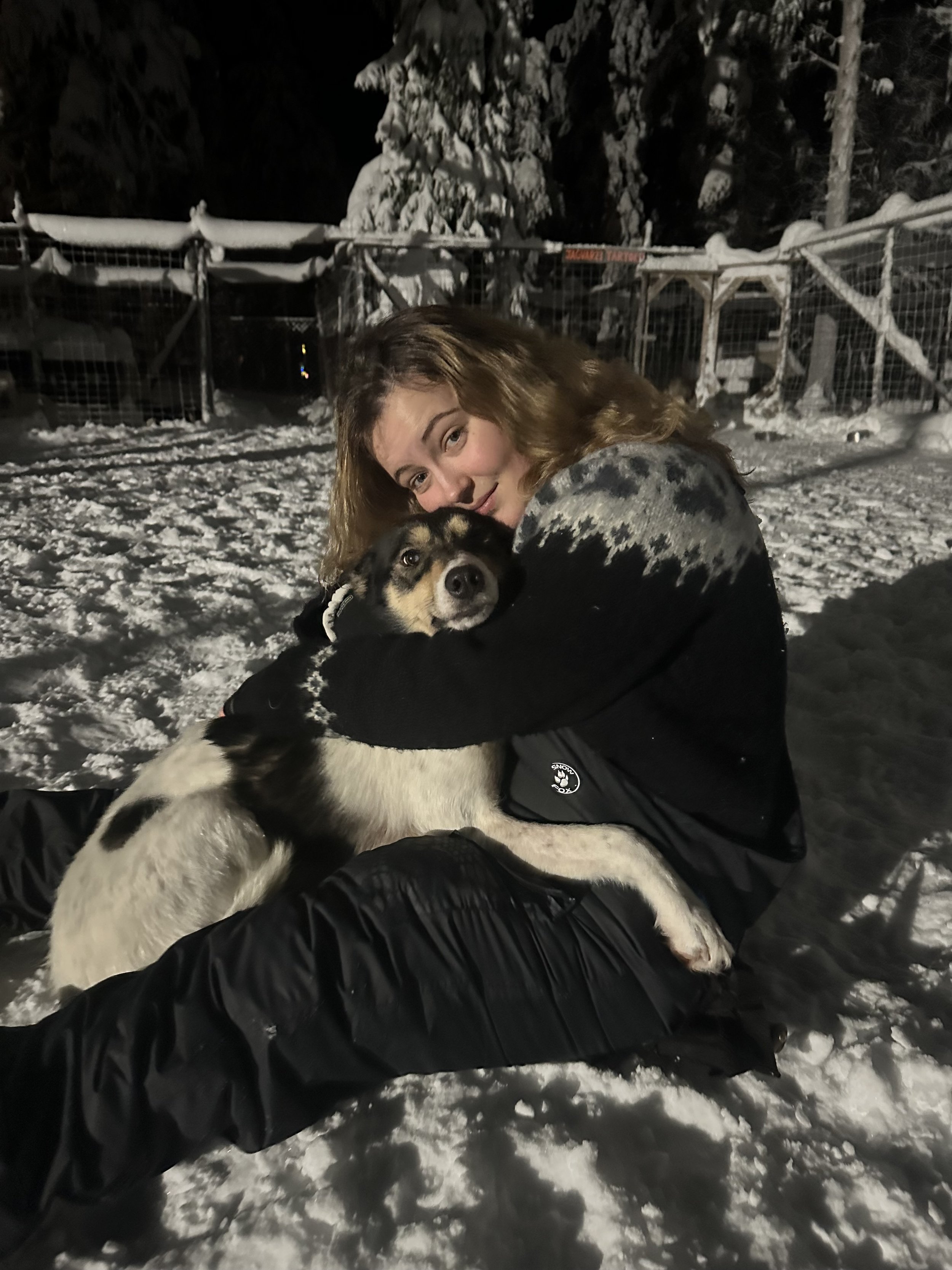 A young woman with light brown curly hair hugging a black and white dog in a snowy outdoor setting at night.