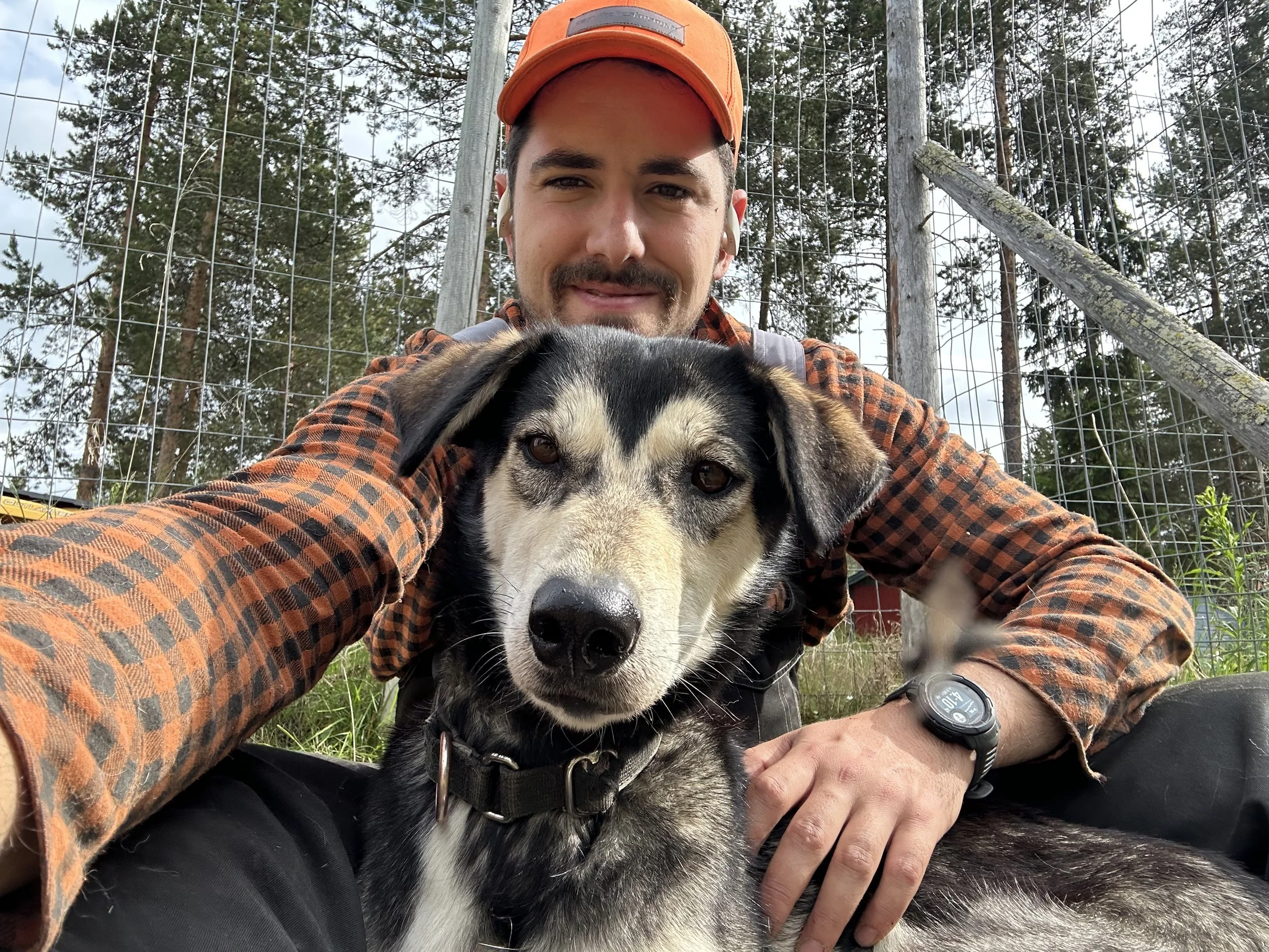A man in an orange cap and plaid shirt taking a selfie with a black and white dog in front of a wooden fence with trees in the background.