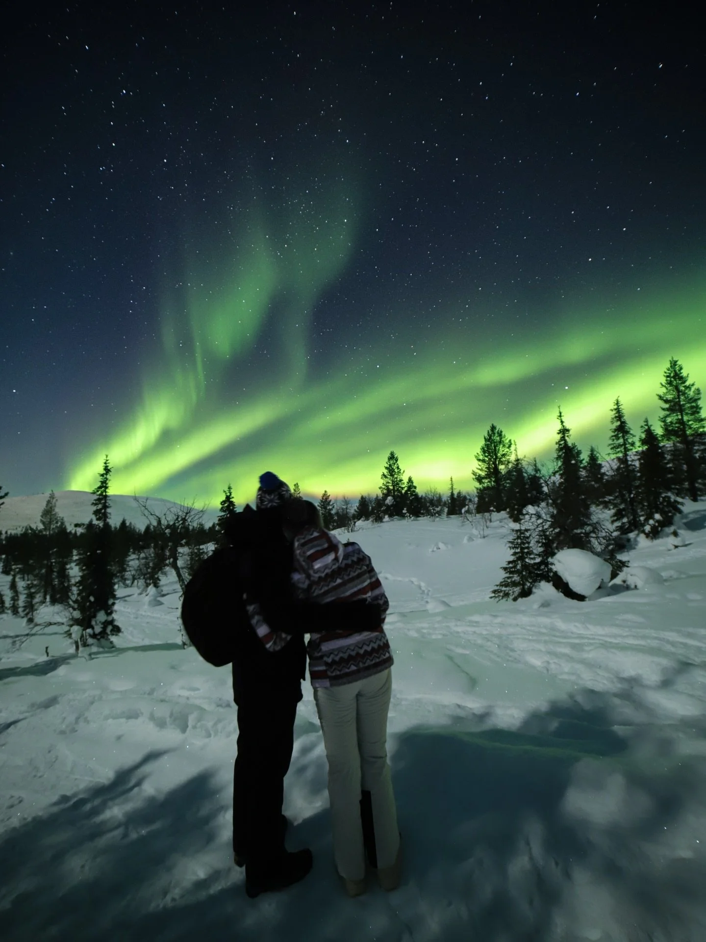 Two people standing in snow, embracing, and watching the northern lights in the night sky over a forested landscape.