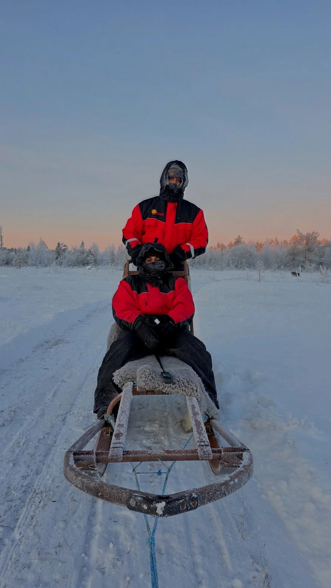 Two people in red and black winter jackets sitting on a sled in a snowy landscape during sunset.