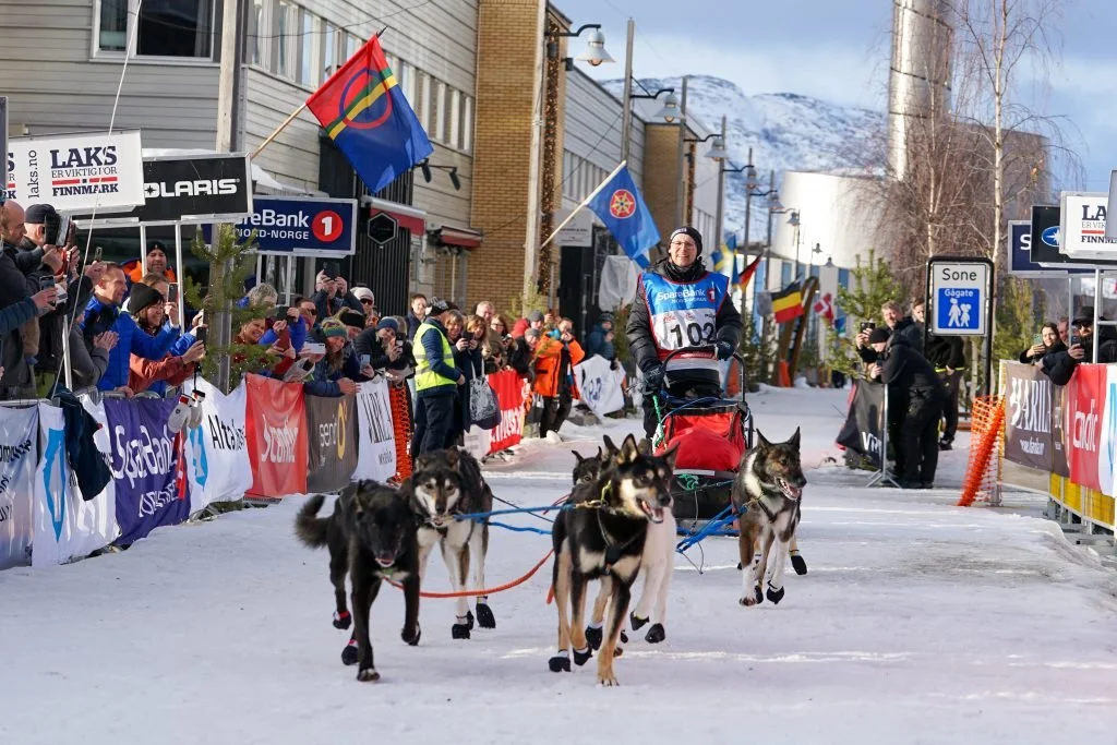 A person is riding a dog sled with four huskies during a winter race, with a crowd of spectators cheering along a snowy street.