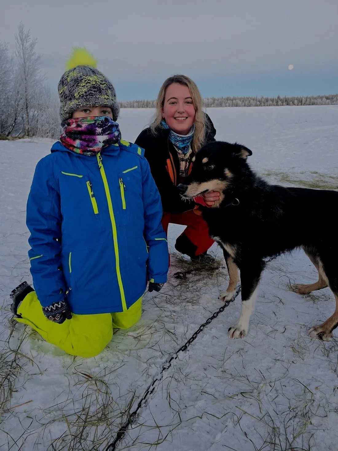 A woman and a young child outdoors in a snowy landscape with a dog, wearing winter clothing. The woman is kneeling, smiling, while the child is standing bundled up. The dog is leash-bound and has black and white fur.