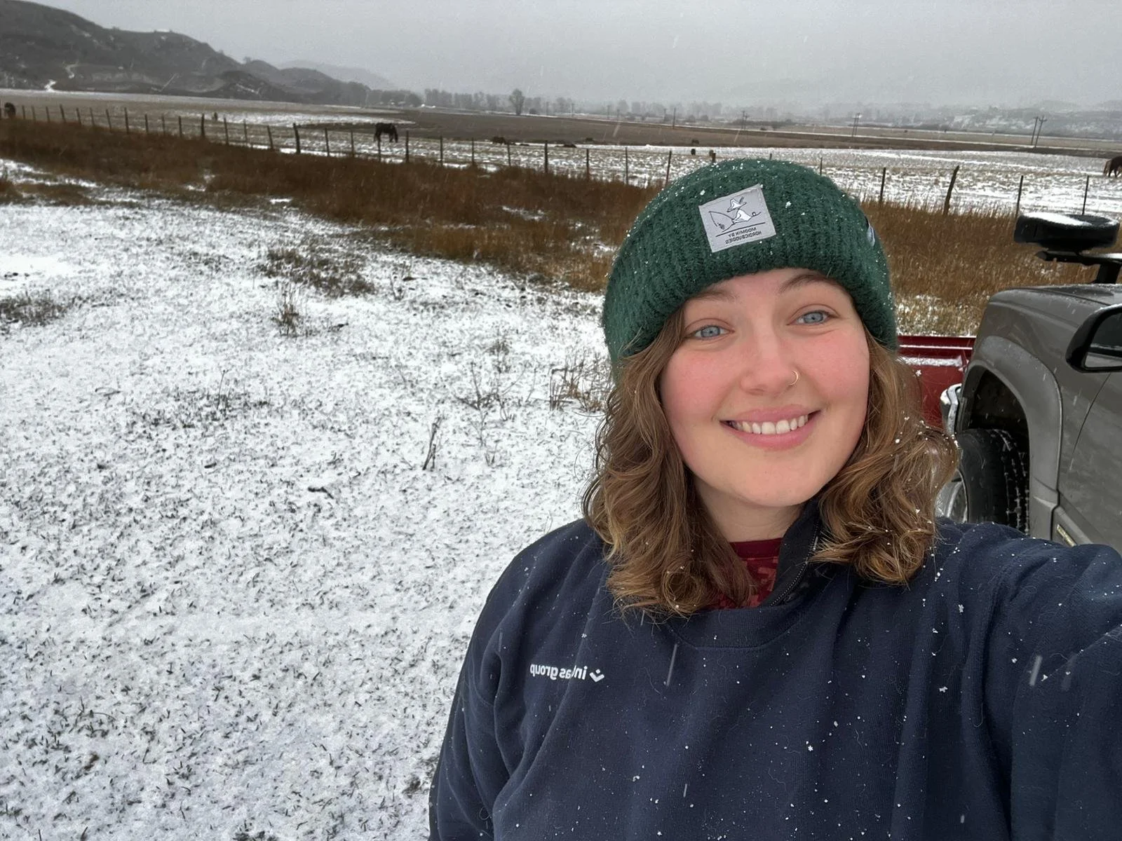 A smiling woman with wavy brown hair, wearing a green knit beanie and a dark jacket, is taking a selfie outdoors in a snow-covered field with a cloudy sky. In the background, there are horses grazing near a fence and mountain ranges in the distance.