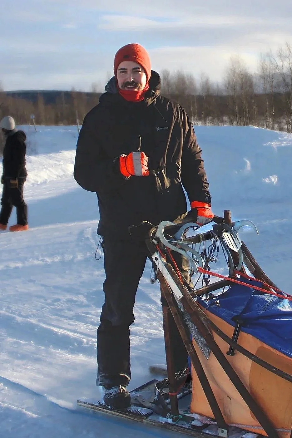 Man in winter clothing standing next to a sled in a snowy landscape, with another person in the background.