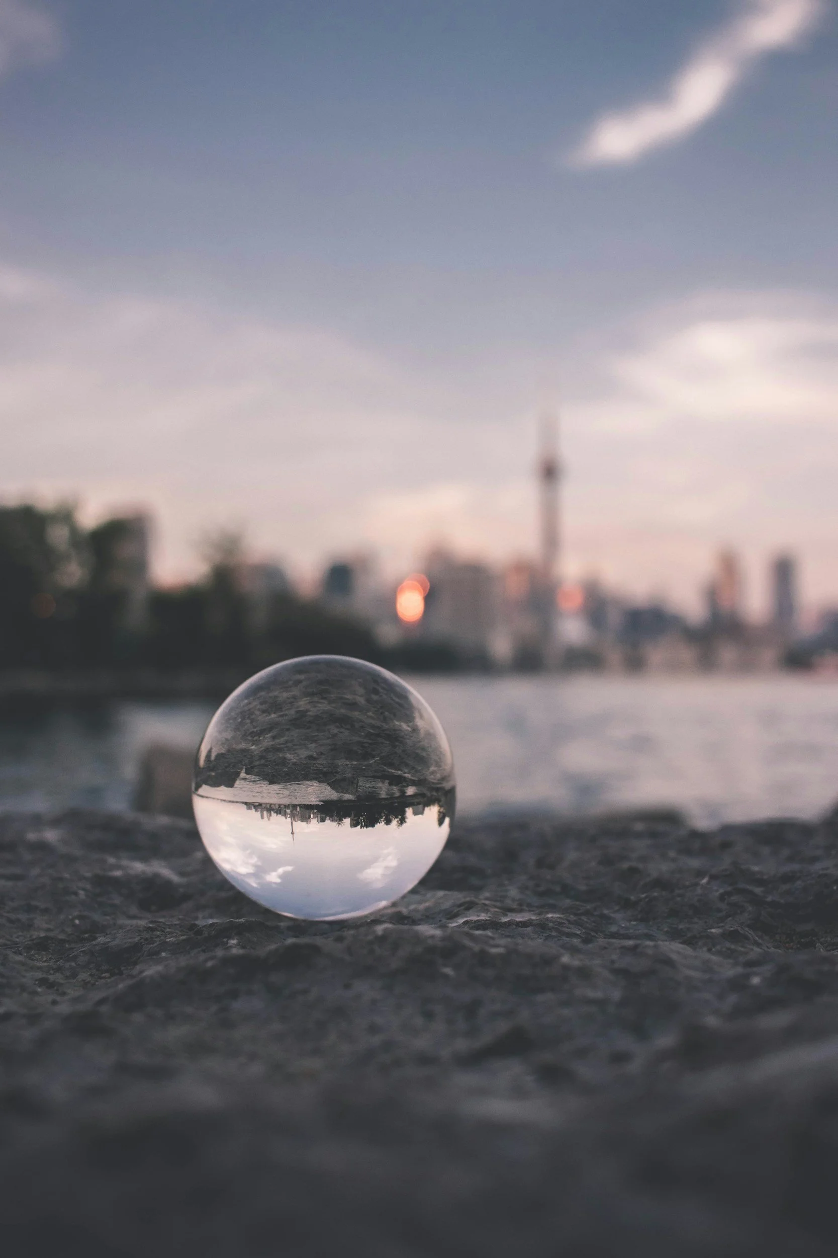 A close-up of a glass ball on a textured surface, with a city skyline and cloudy sky in the background, during late evening or dusk.