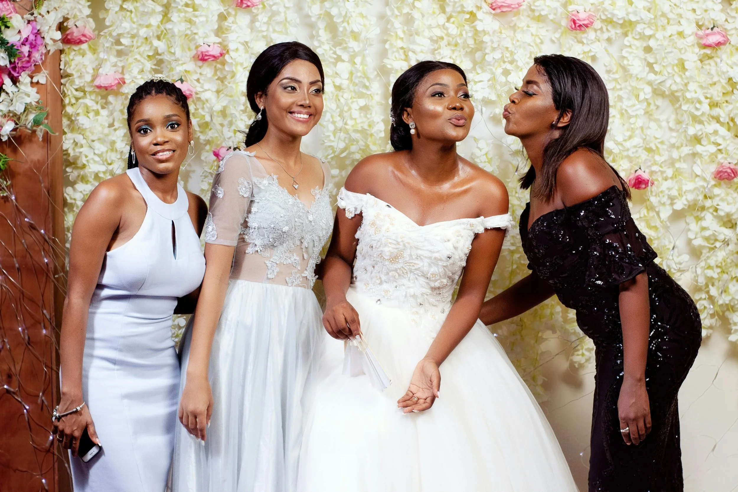 Four women posing at a wedding; one is the bride in a white dress, two are in light-colored dresses, and one in a black dress against a floral backdrop.