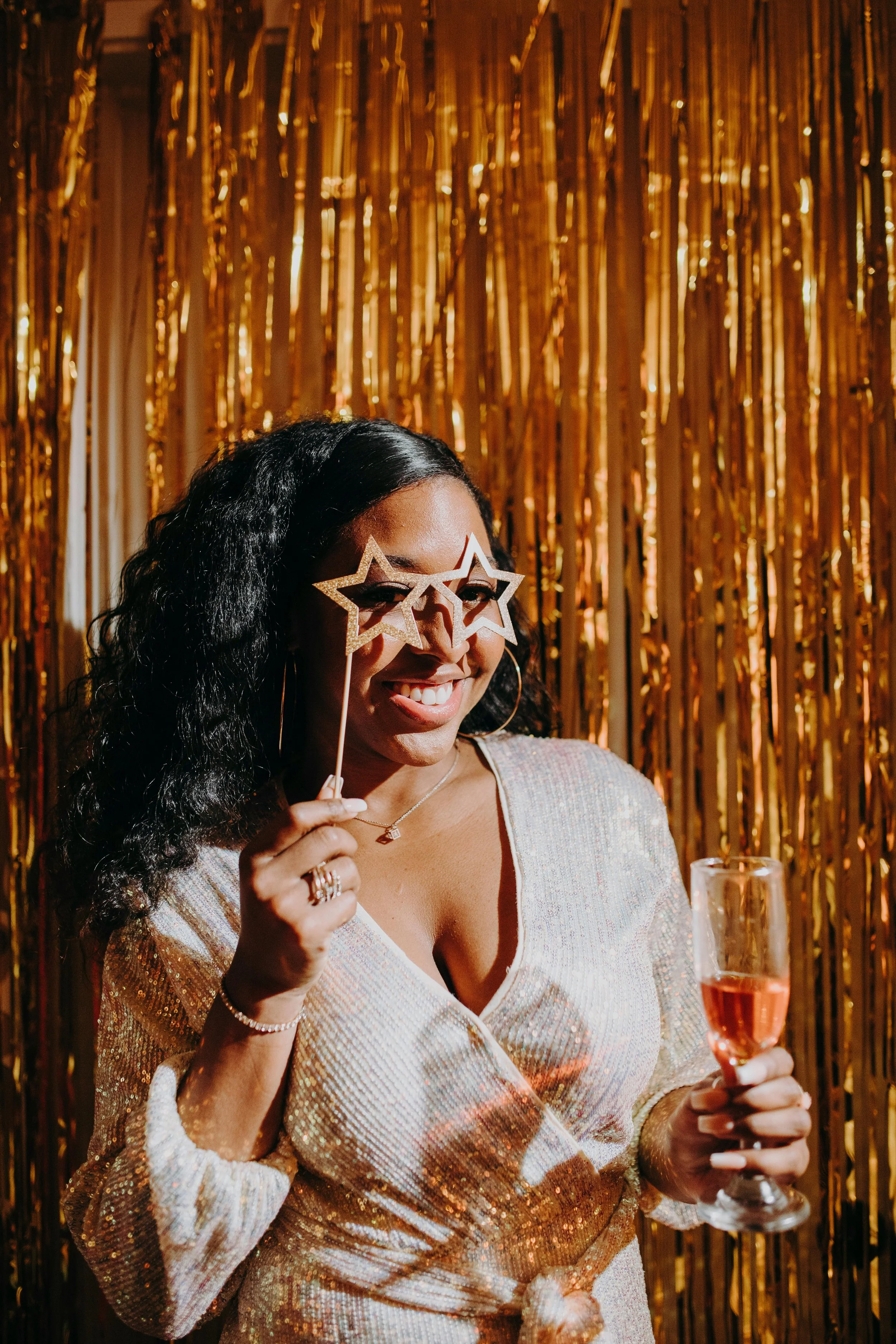 Woman smiling in party attire holding star-shaped glasses and a champagne glass in front of a gold curtain.