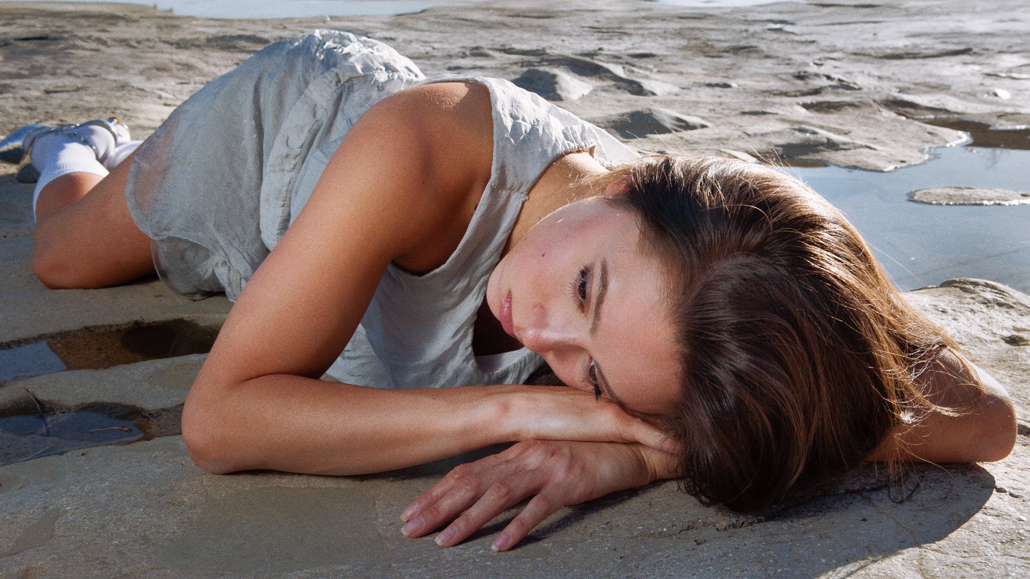 A woman lying on her side on a rocky surface near a body of water, with her eyes closed and her head resting on her arm.