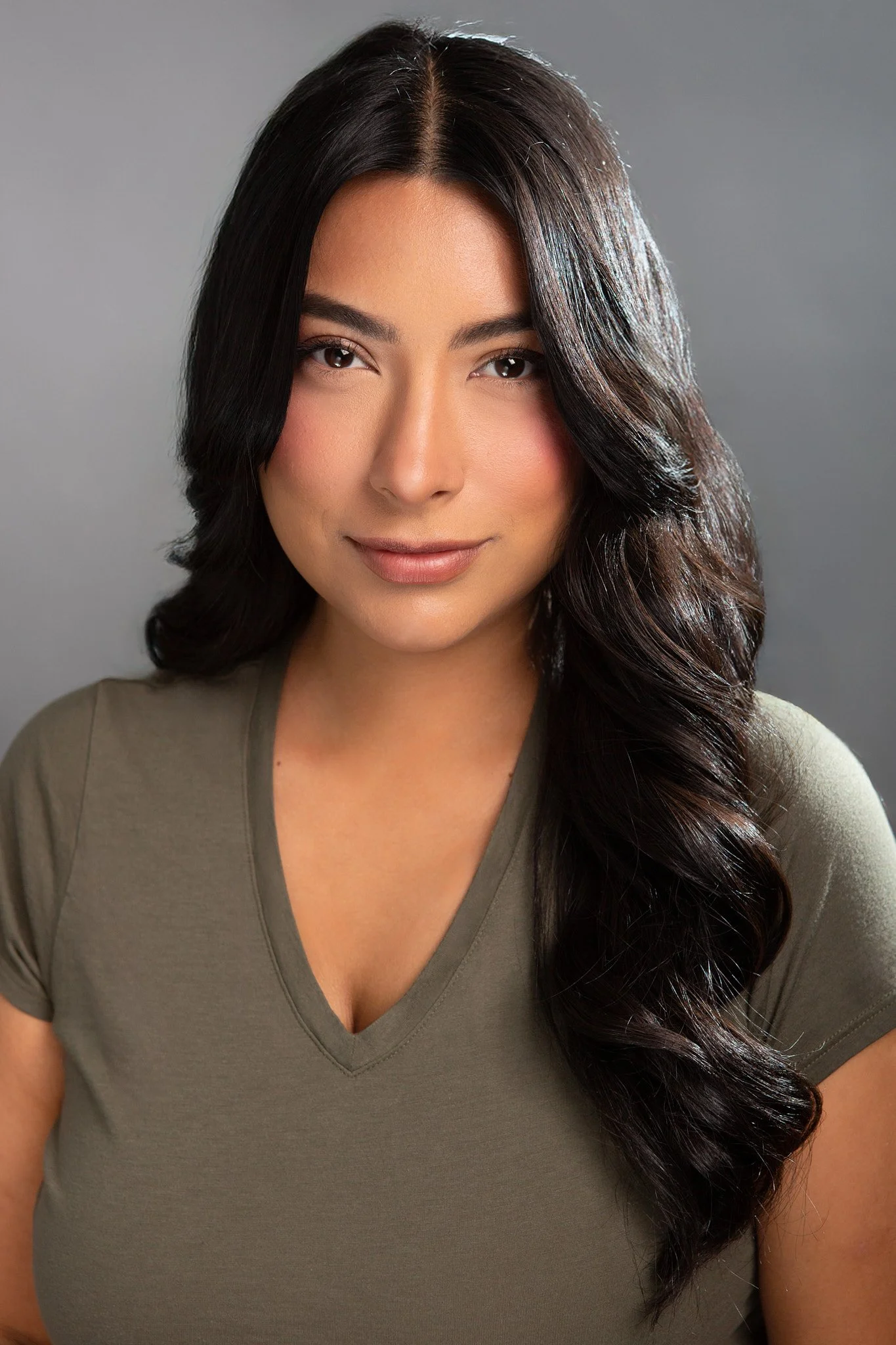 A woman with long, wavy dark hair, wearing a khaki V-neck T-shirt, looking directly at the camera against a gray background.