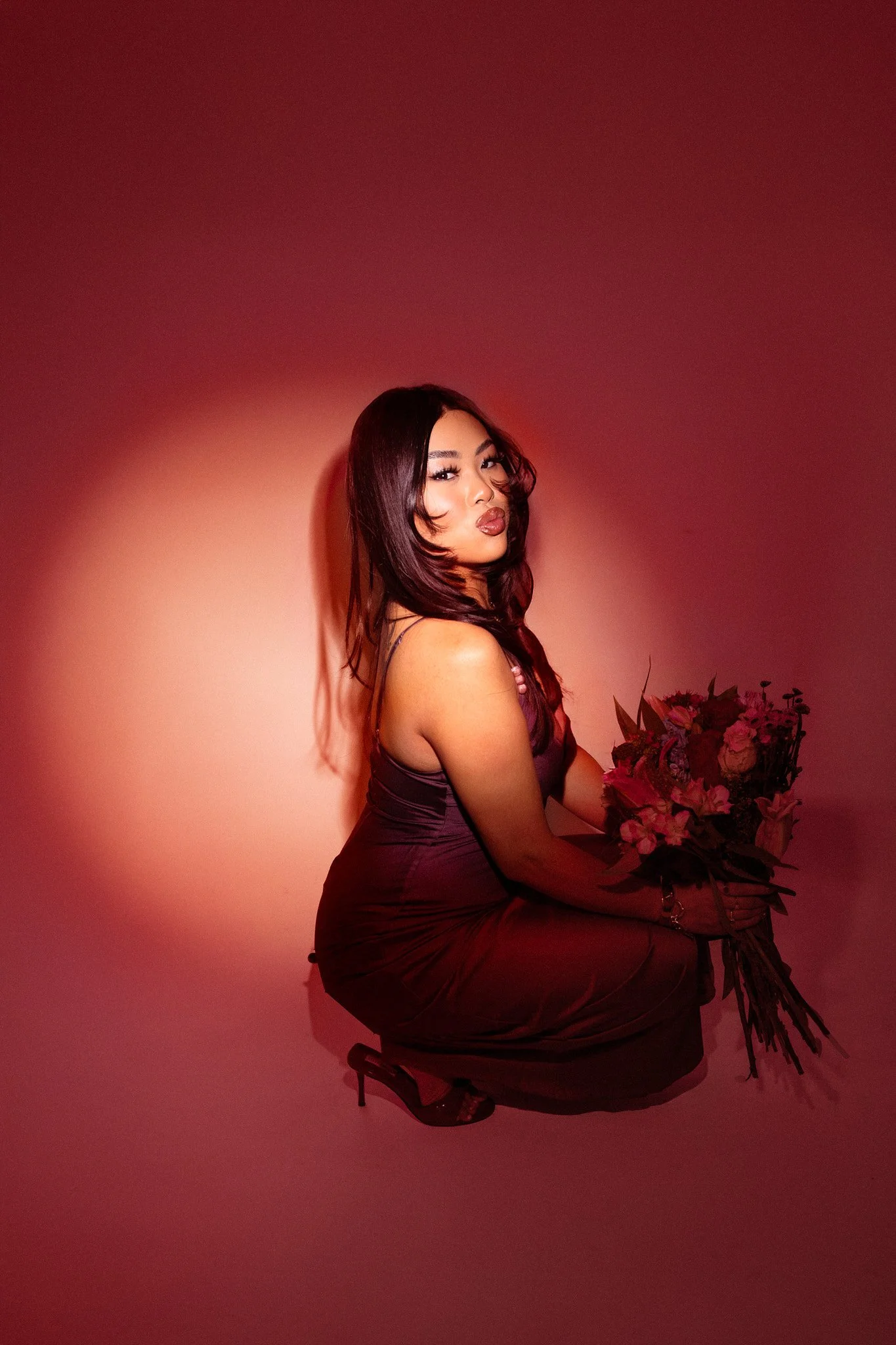 A woman with long dark hair wearing a purple dress and high heels, kneeling down and holding a bouquet of flowers, posing against a pink gradient background.