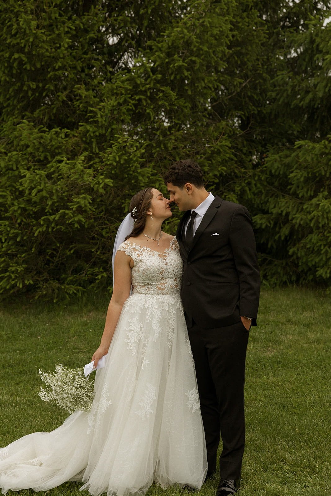 A bride and groom stand close together outdoors, touching foreheads, with large green trees in the background. The bride wears a white lace wedding gown with a veil and holds a small bouquet of baby's breath flowers. The groom is dressed in a black s