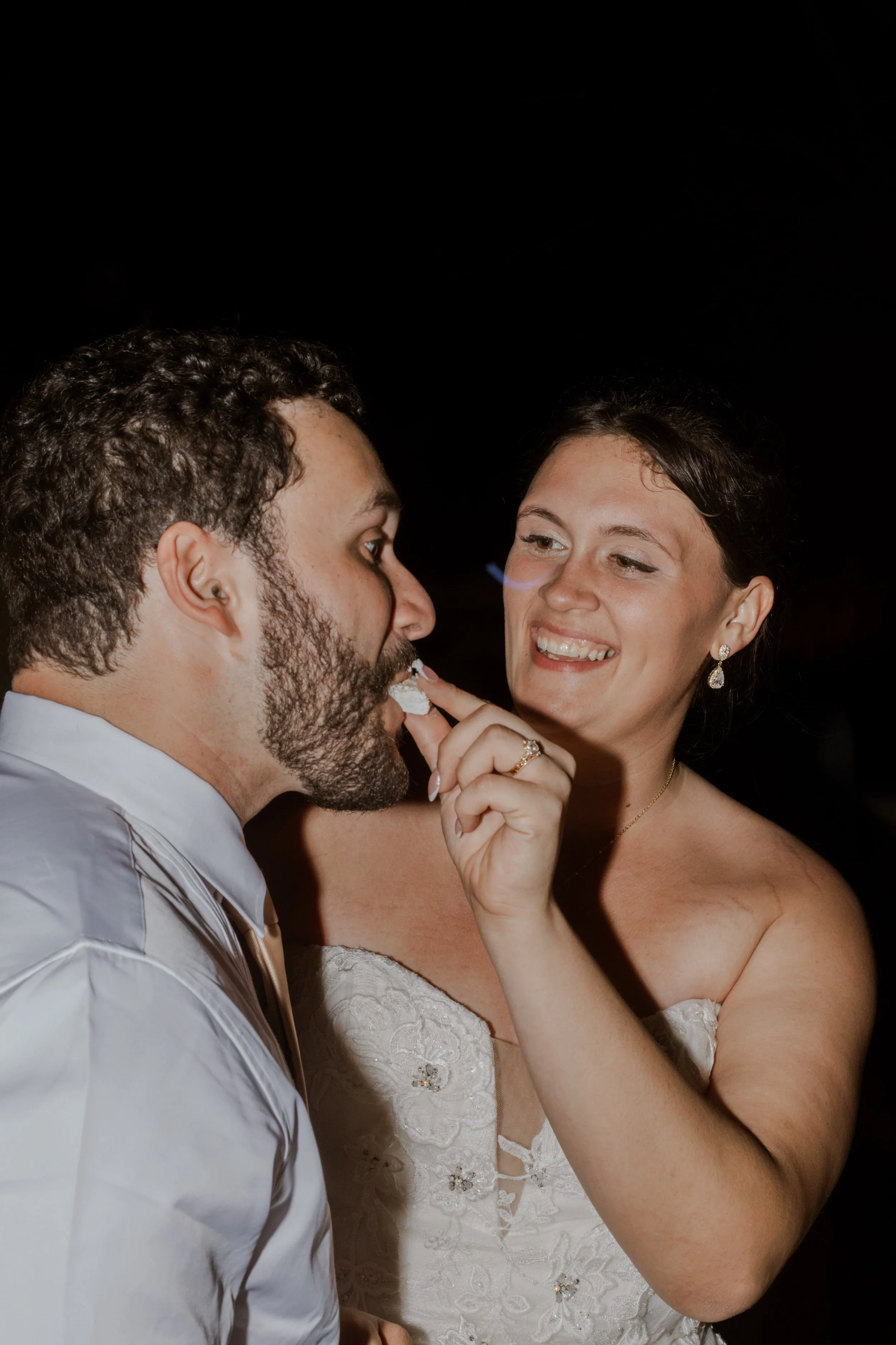 A brunette woman in a strapless wedding dress feeding her husband cake at their wedding reception