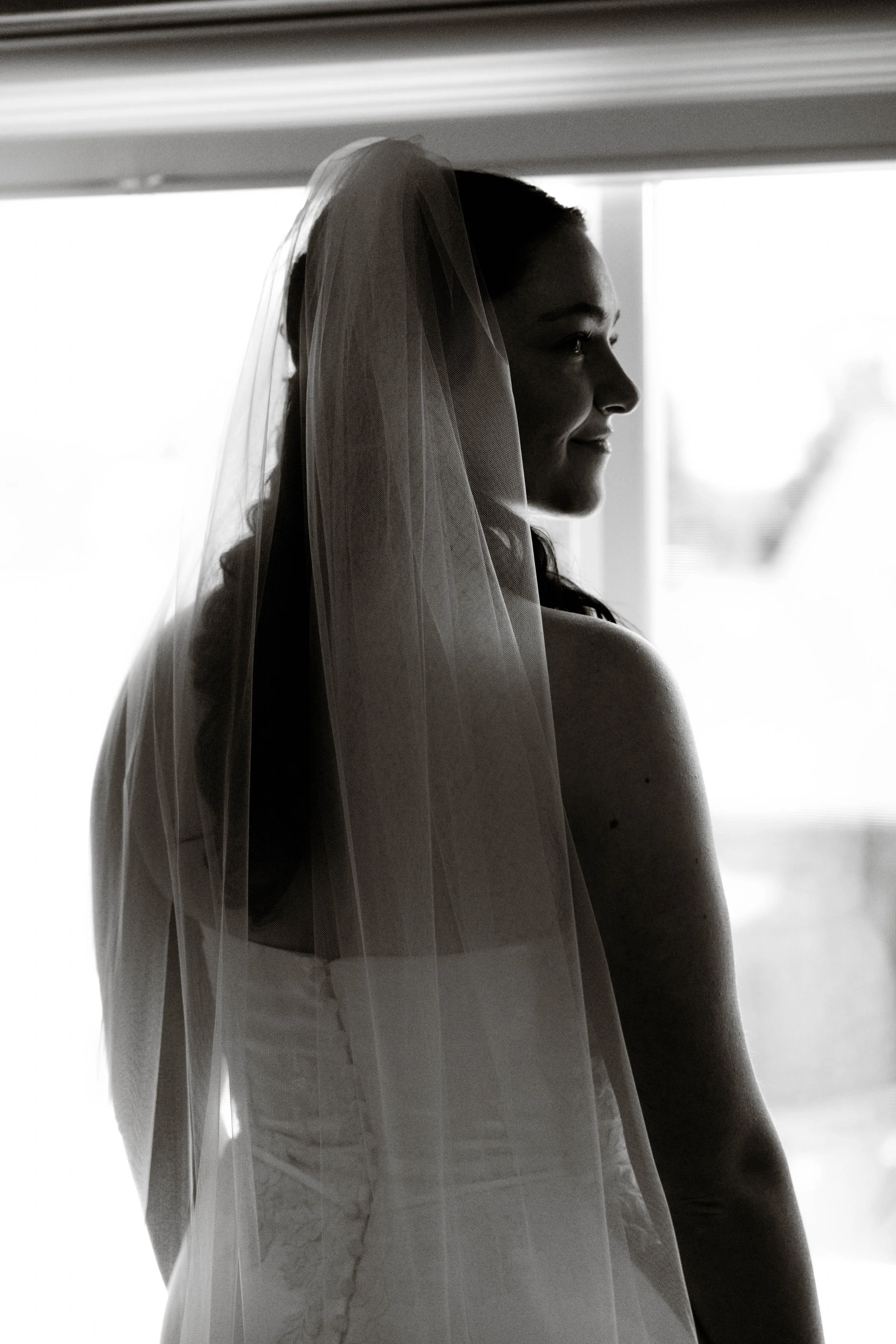 Black and white bridal portrait photography of a bride wearing a wedding veil, standing by a window in soft natural light during wedding day preparations.