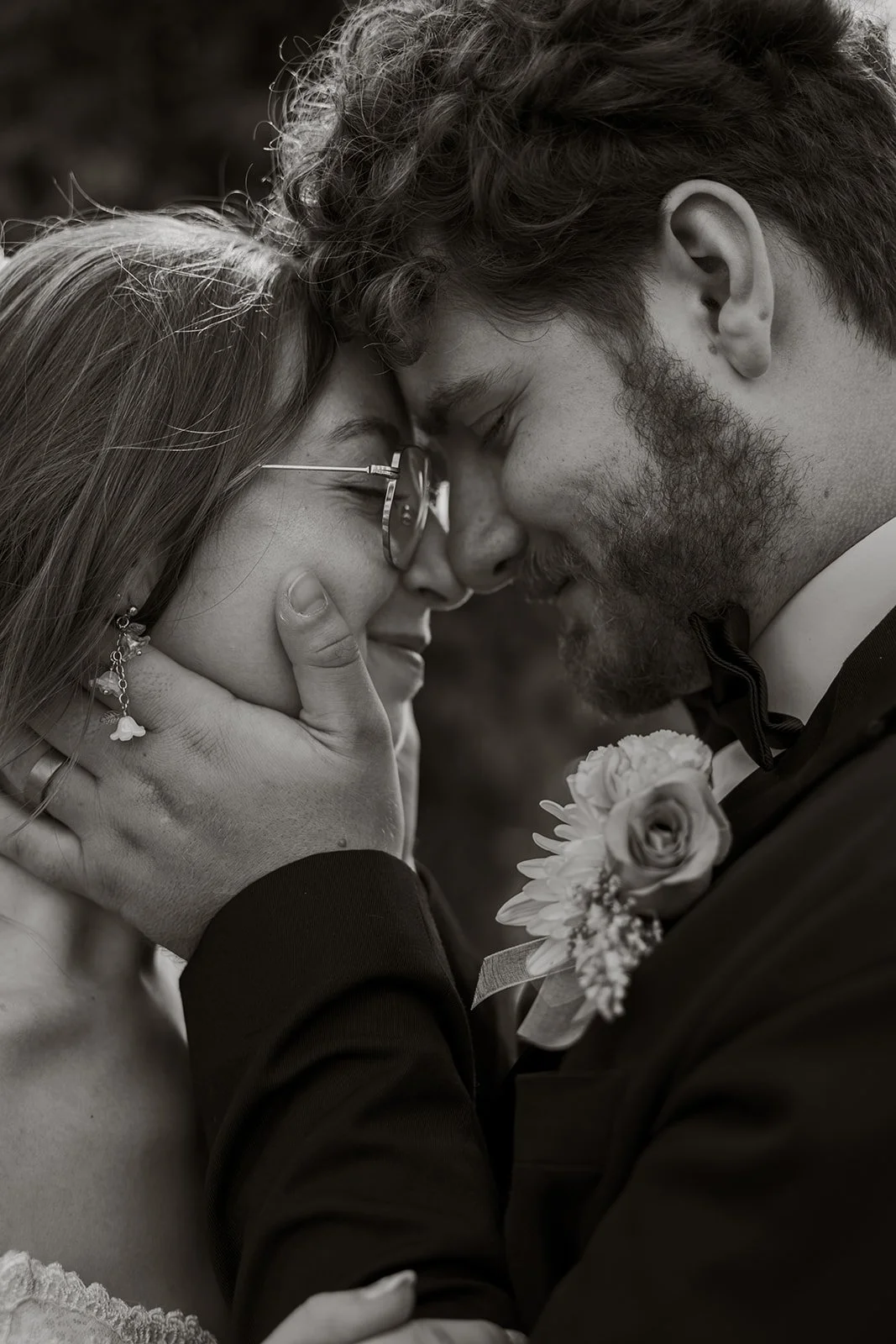 A black and white photo of a couple with their foreheads touching, eyes closed, sharing an intimate moment. The woman is wearing glasses and earrings, and the man is in a suit with a boutonnière.