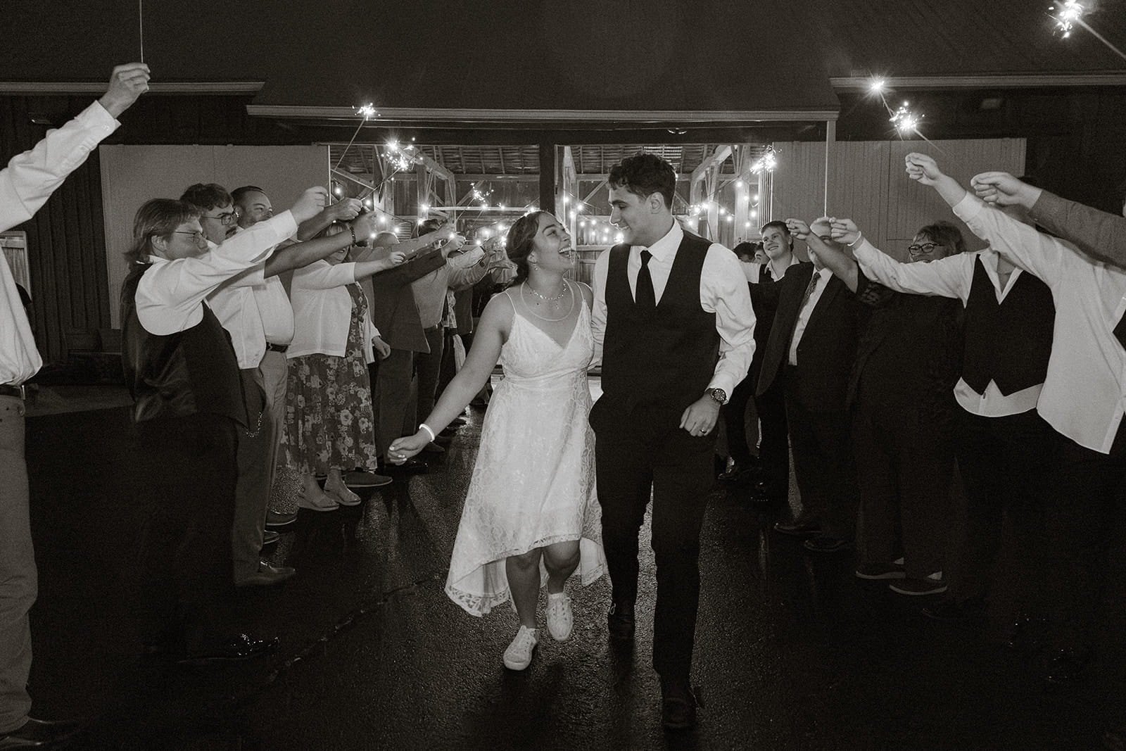 Black-and-white photo of a bride and groom walking through an aisle of friends holding sparklers at night.