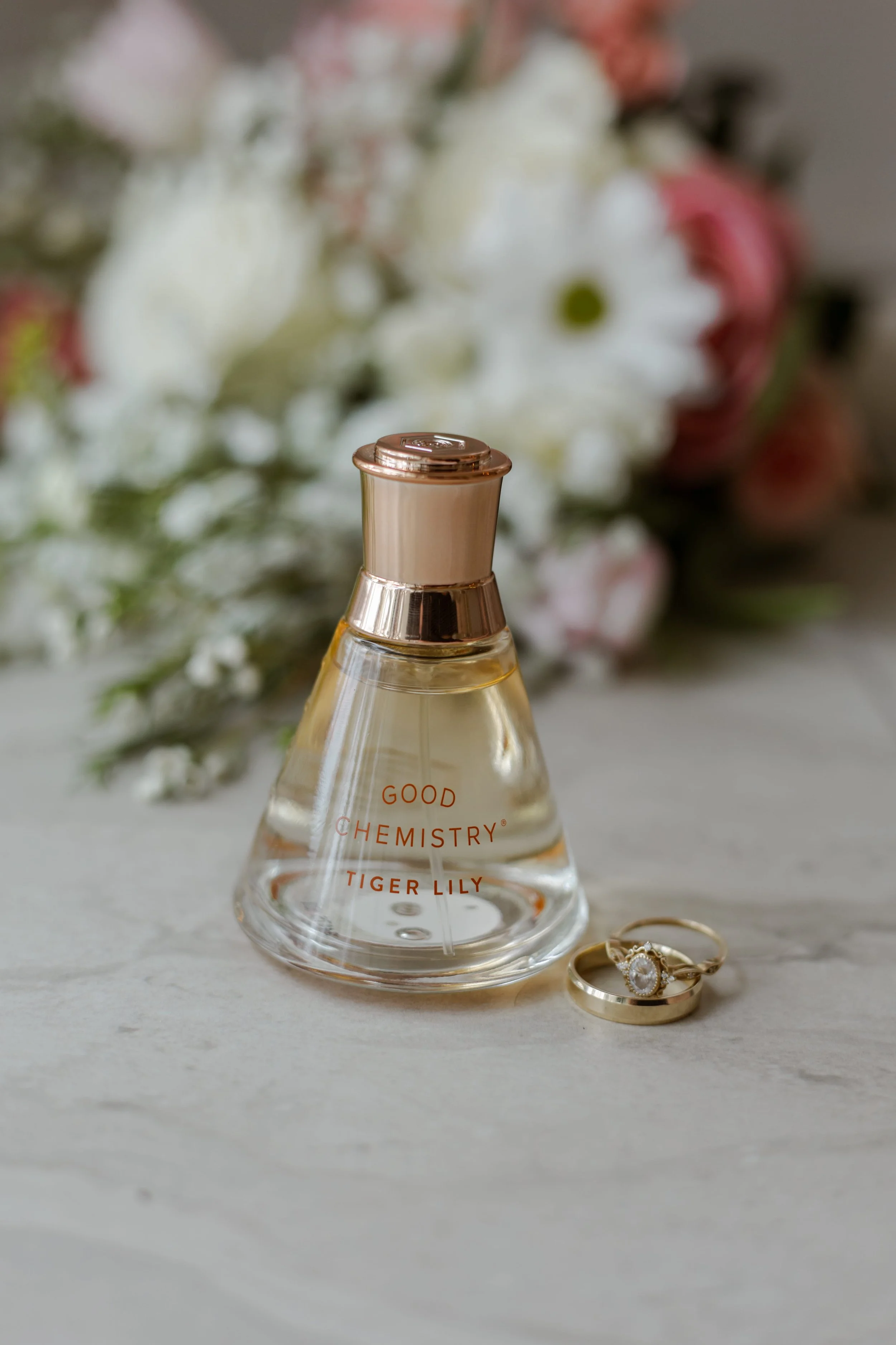 Wedding detail photography of bridal perfume, engagement ring, and wedding band styled together on a neutral surface, captured during wedding day getting ready details.