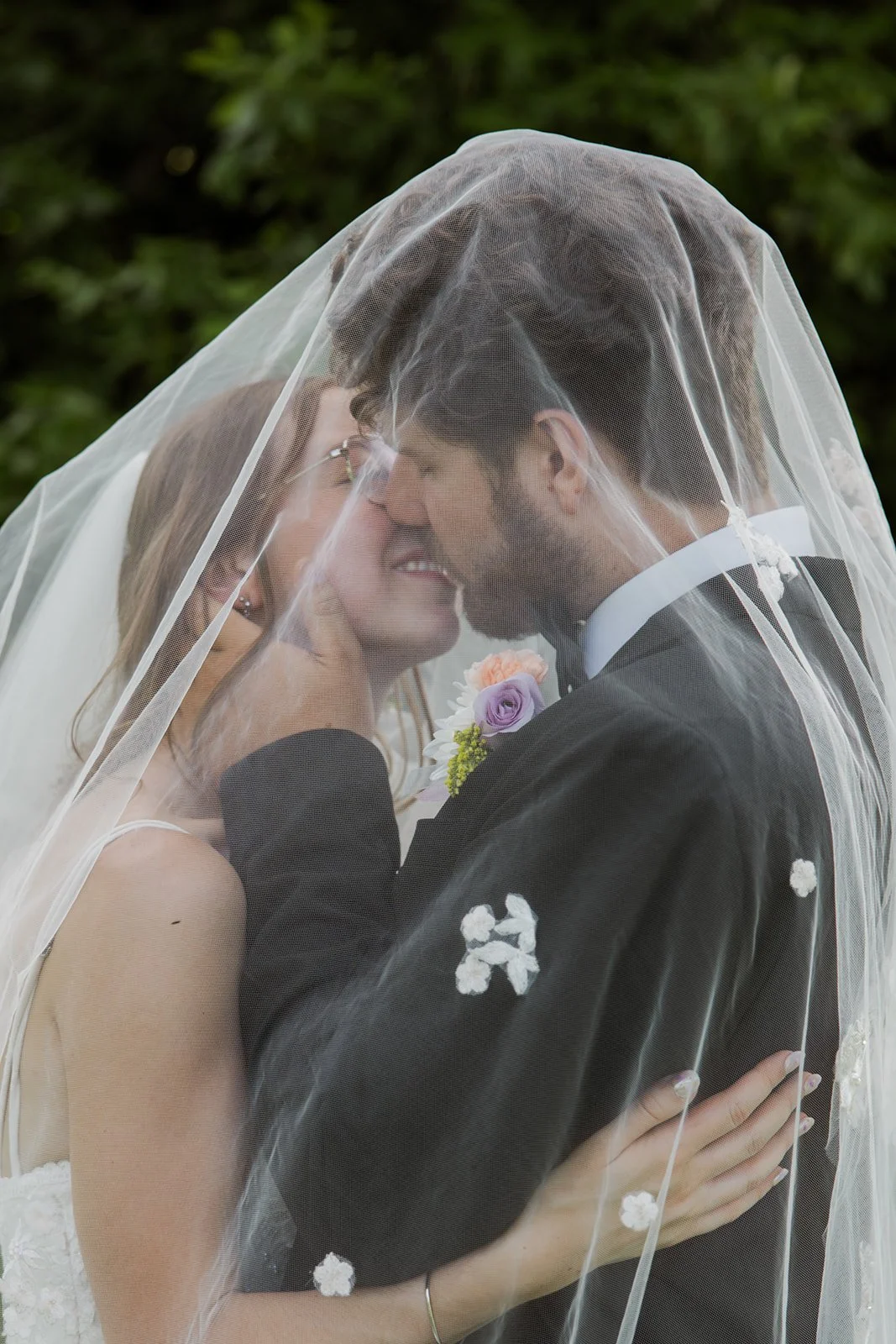 A bride and groom sharing a kiss under a wedding veil, with greenery in the background.