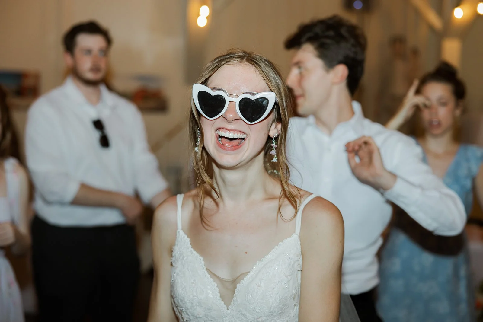A woman wearing large heart-shaped sunglasses smiles and laughs at a party or celebration with other people in the background.