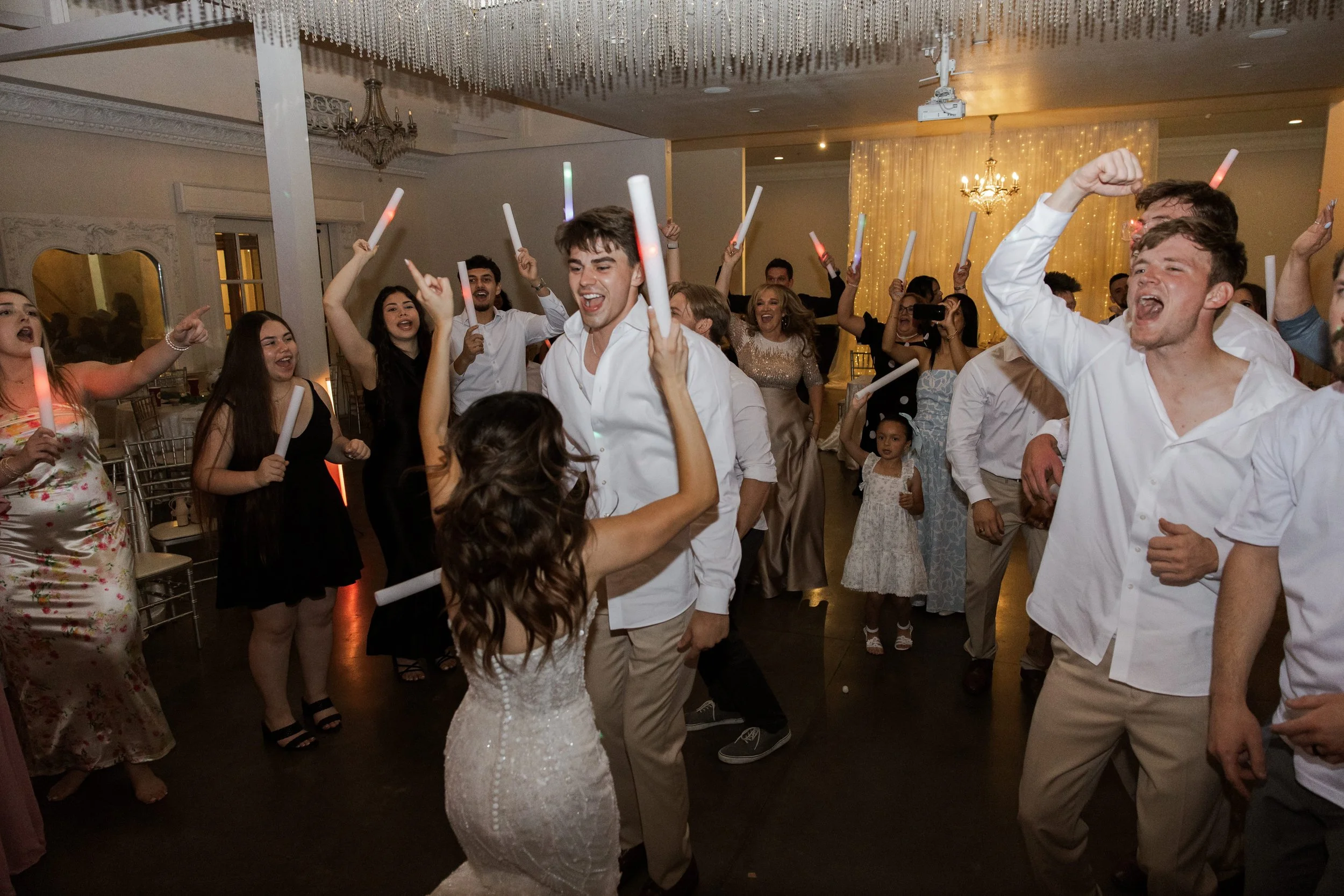 People dancing and celebrating at a wedding reception, with some holding glow sticks and smiling.