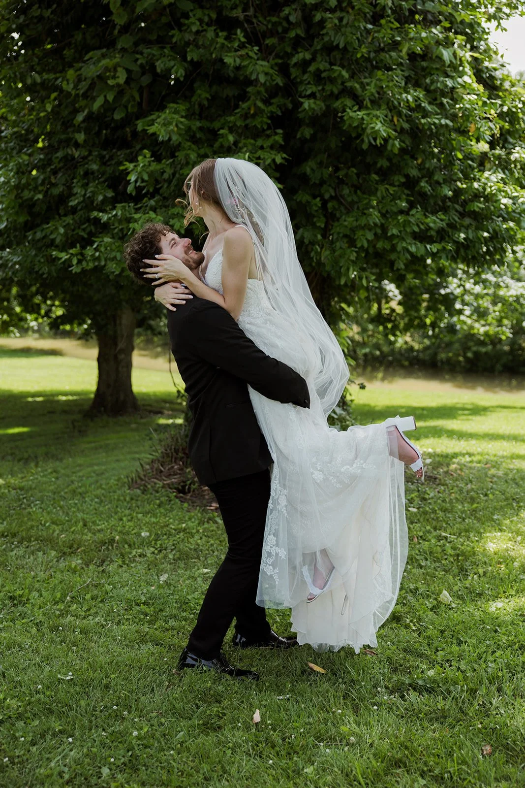 A groom lifting a bride outdoors with green grass and trees in the background.
