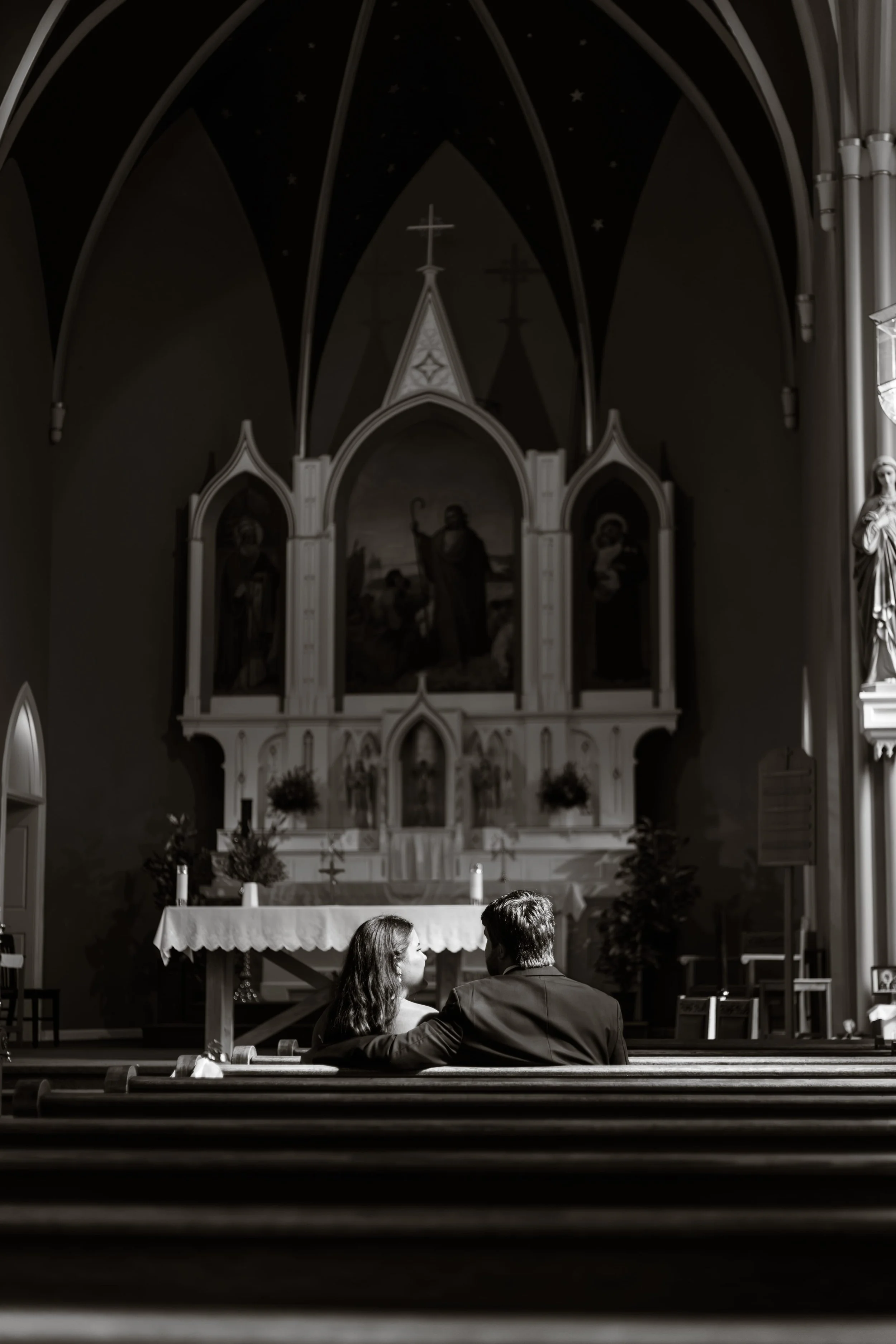 Black and white wedding photography of a bride and groom sitting together on a church pew, sharing a quiet, intimate moment before their ceremony inside a traditional church wedding venue.