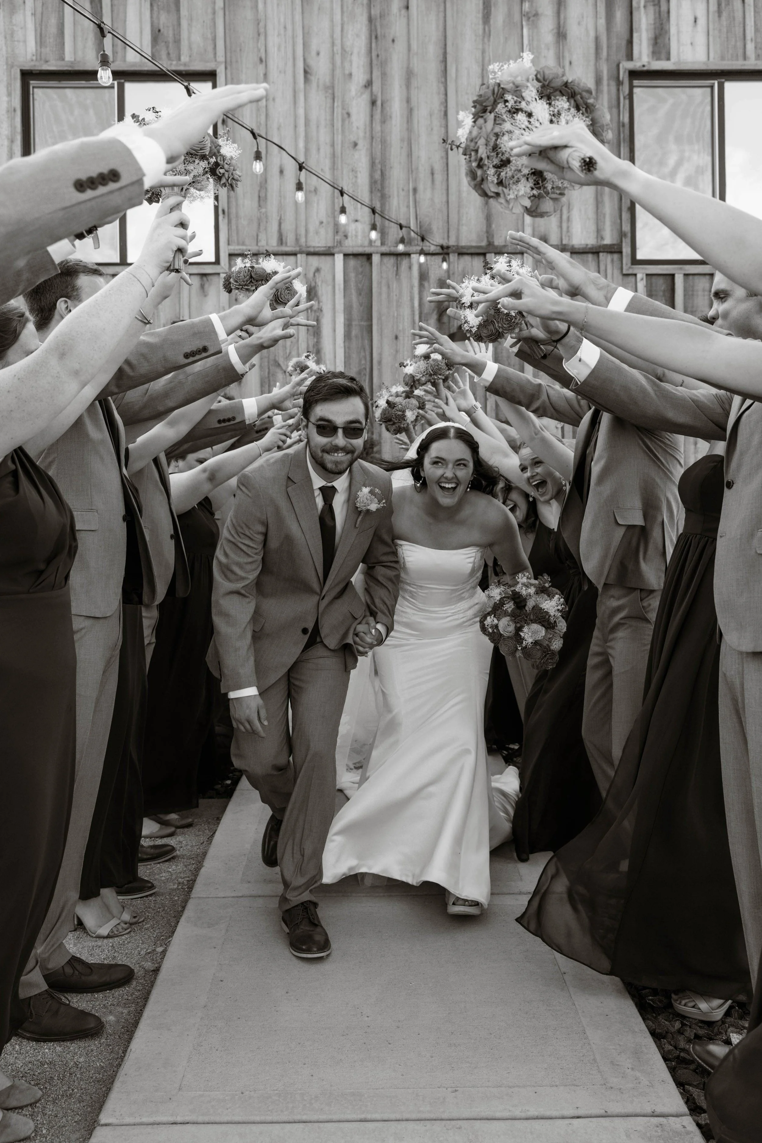 A joyous bride and groom walking through a tunnel of friends and family holding bouquets and throwing flower petals at a wedding ceremony.