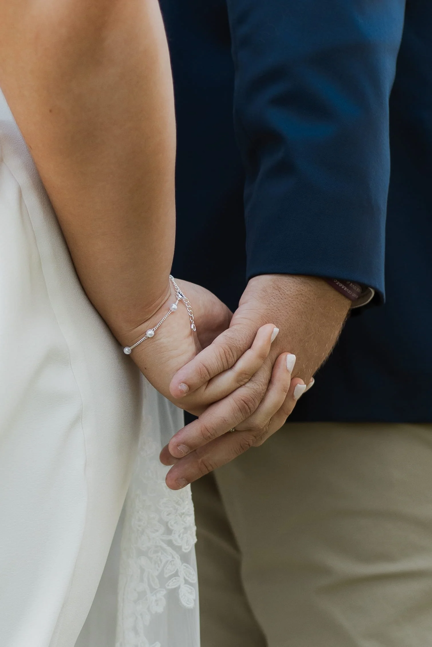 Wedding photography detail of a bride and groom holding hands, showing wedding rings and bridal bracelet during an intimate wedding day moment.