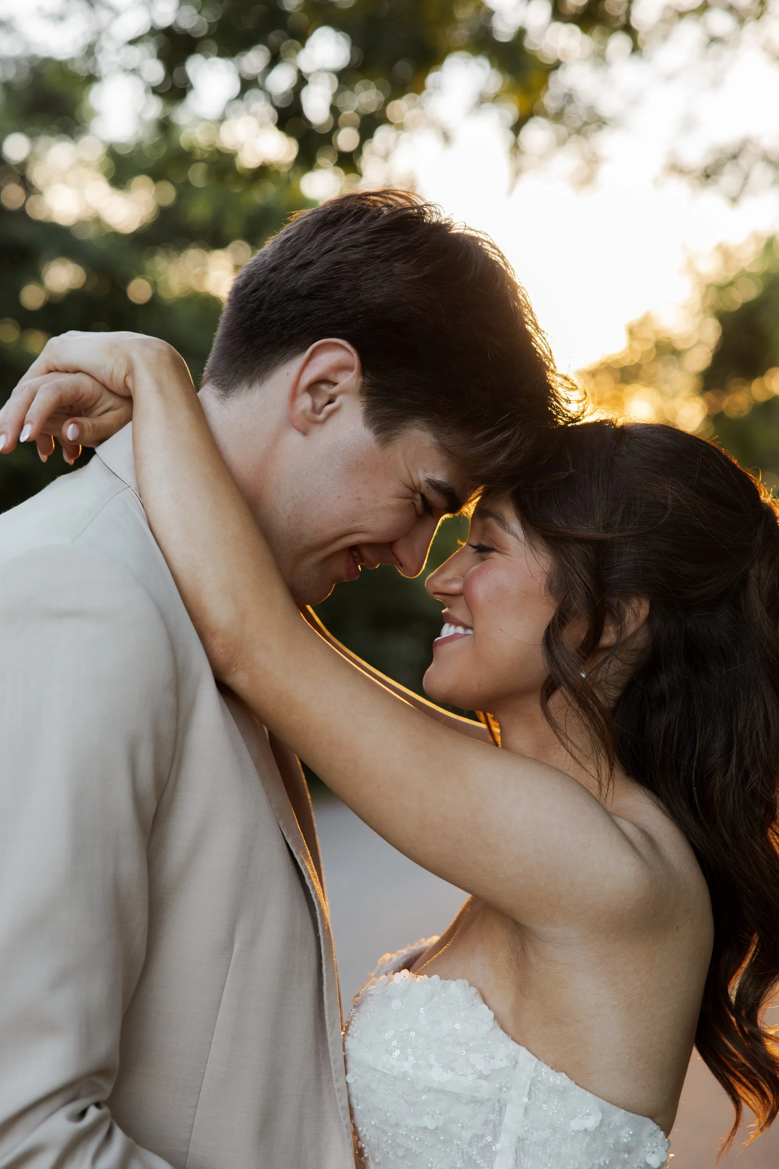 A couple dressed in wedding attire embracing in a romantic moment outdoors during sunset, with trees and sunlight in the background.