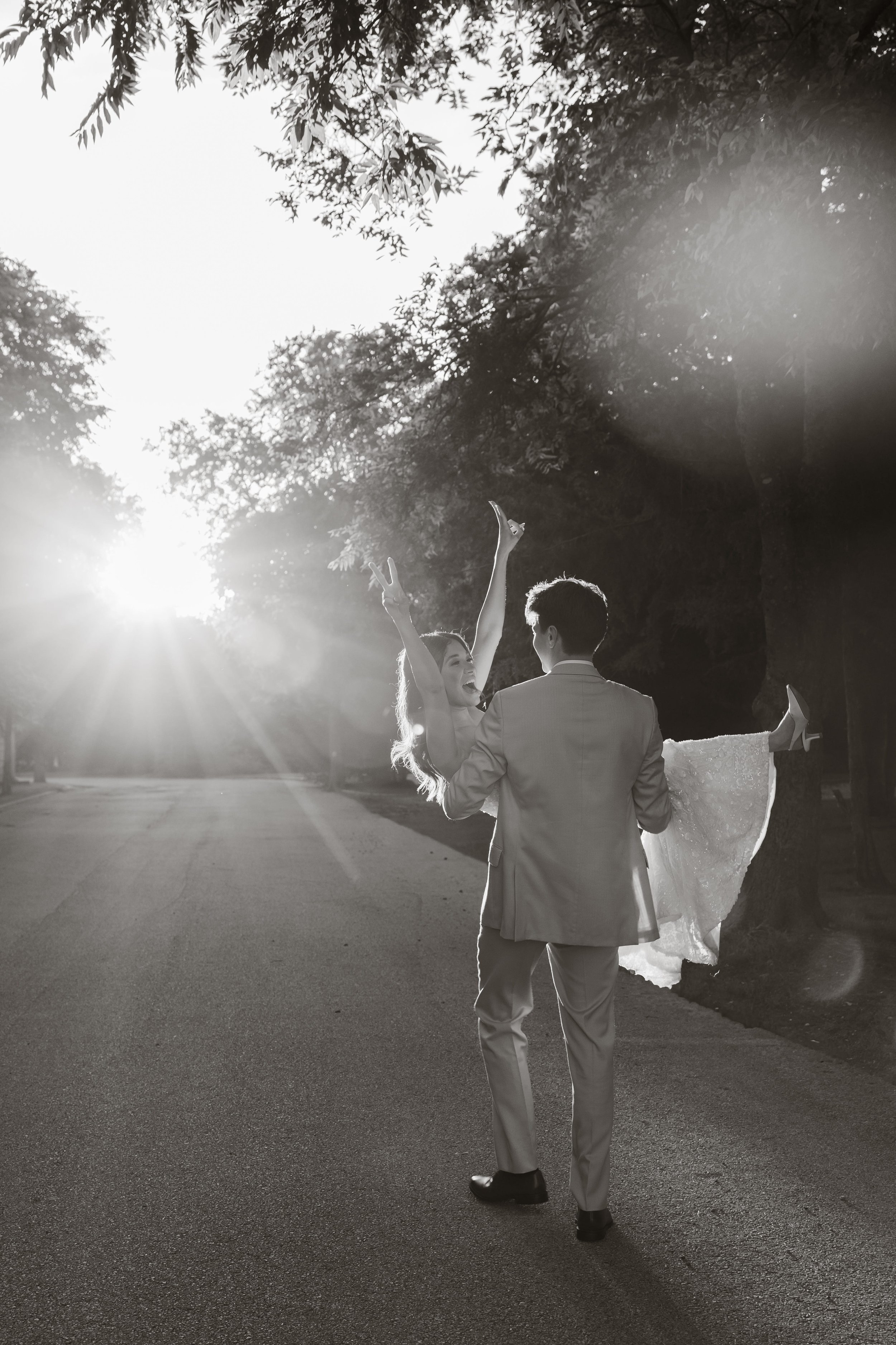 A couple, dressed in wedding attire, is dancing on a street with sunlight filtering through trees in the background. The woman is lifting her arms and smiling, while the man faces her.