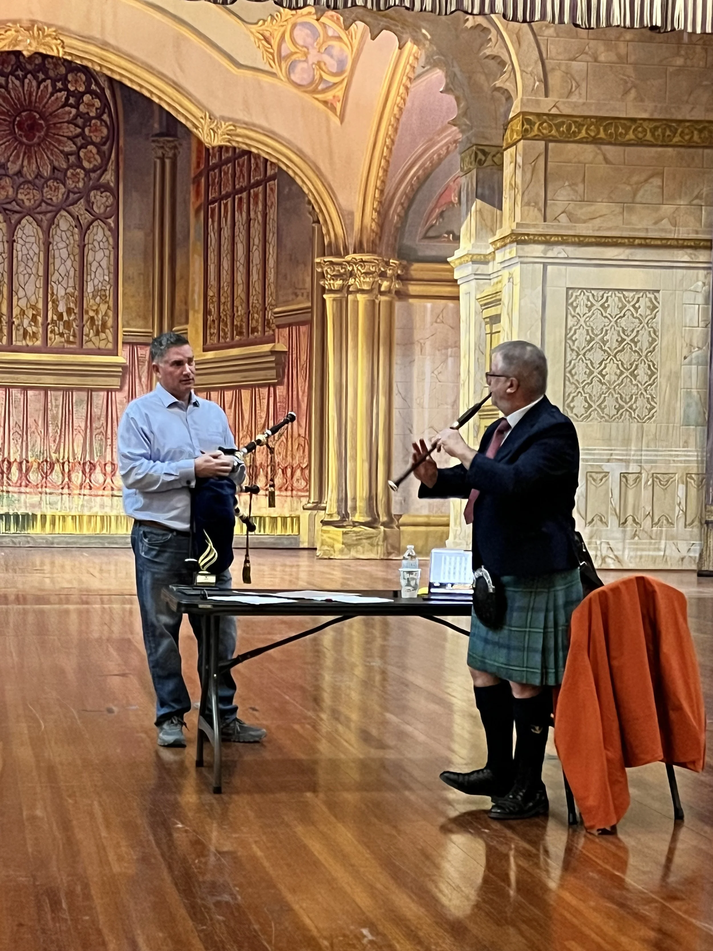 Two men playing bagpipes on a stage with ornate, gold-colored, Gothic-style architectural backdrop.