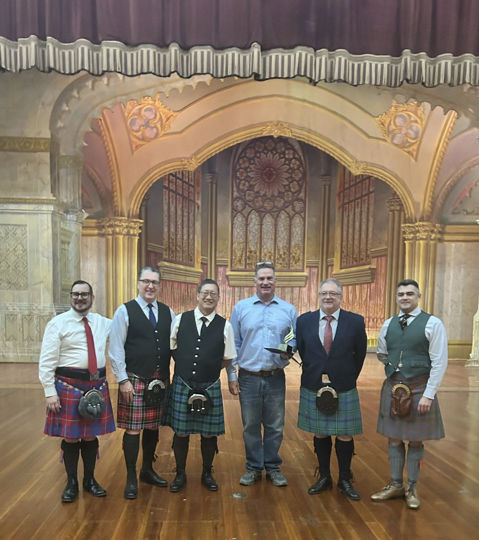 A group of six men standing on a wooden floor, dressed in kilts and formal shirts. Behind them is a stage with ornate, gold-accented architecture and stained glass windows, creating the appearance of a theatrical or concert venue.