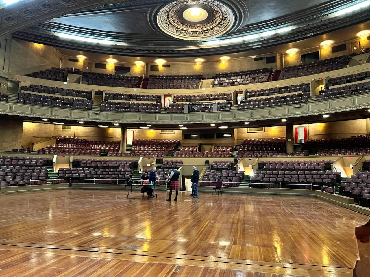 Empty auditorium with a wooden stage, ornate ceiling, and multiple tiers of empty seating. Four people are standing and sitting near the stage.