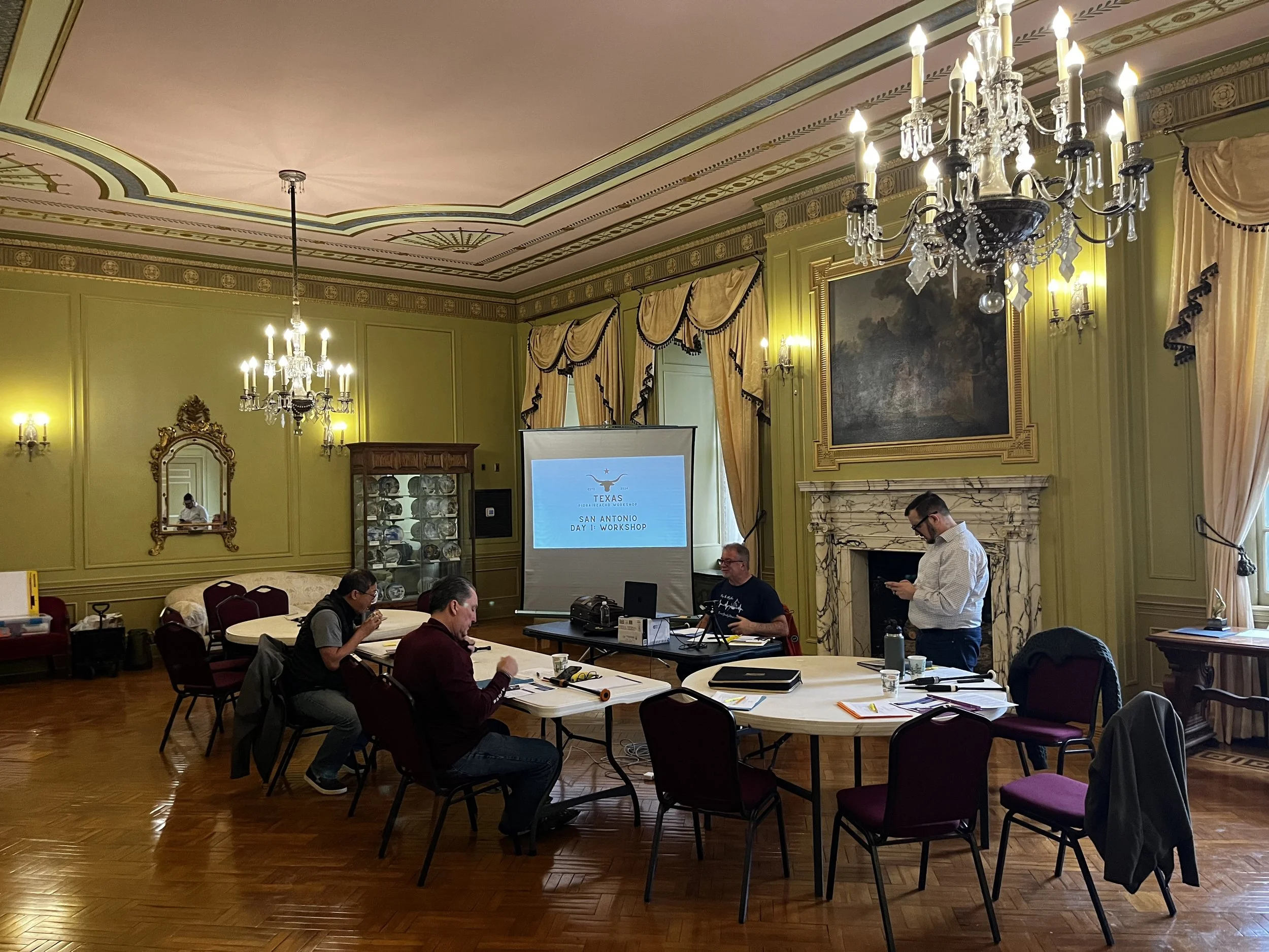 A group of people in a luxurious, ornate conference room with high ceilings, chandeliers, and gold curtains, participating in a workshop or meeting with a presentation screen displaying the Texas flag and text that reads "San Antonio Day 1 Workshop."