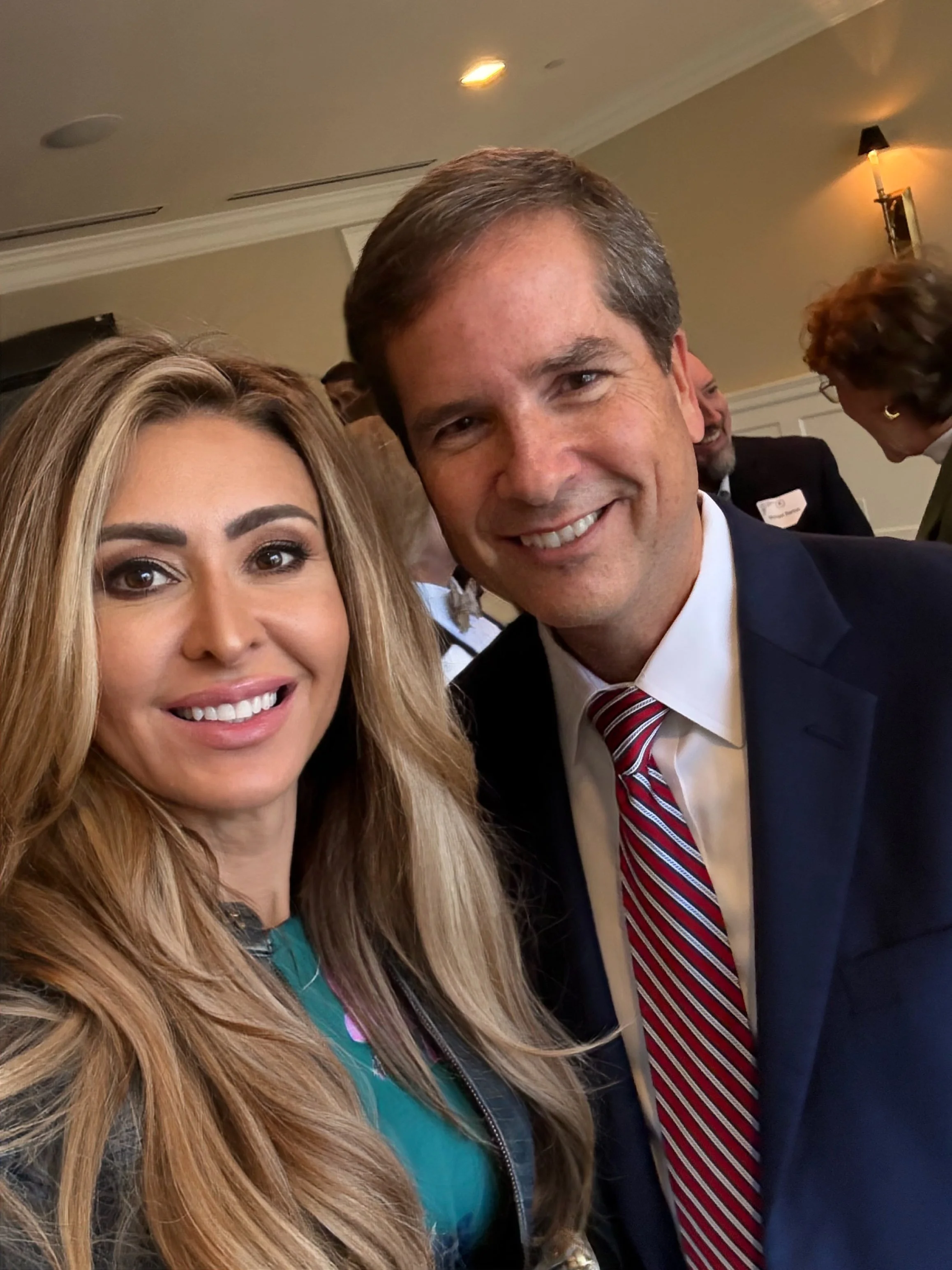 A woman with long, wavy blonde hair taking a selfie with a man in a dark suit and red striped tie at an indoor event, with other attendees in the background.
