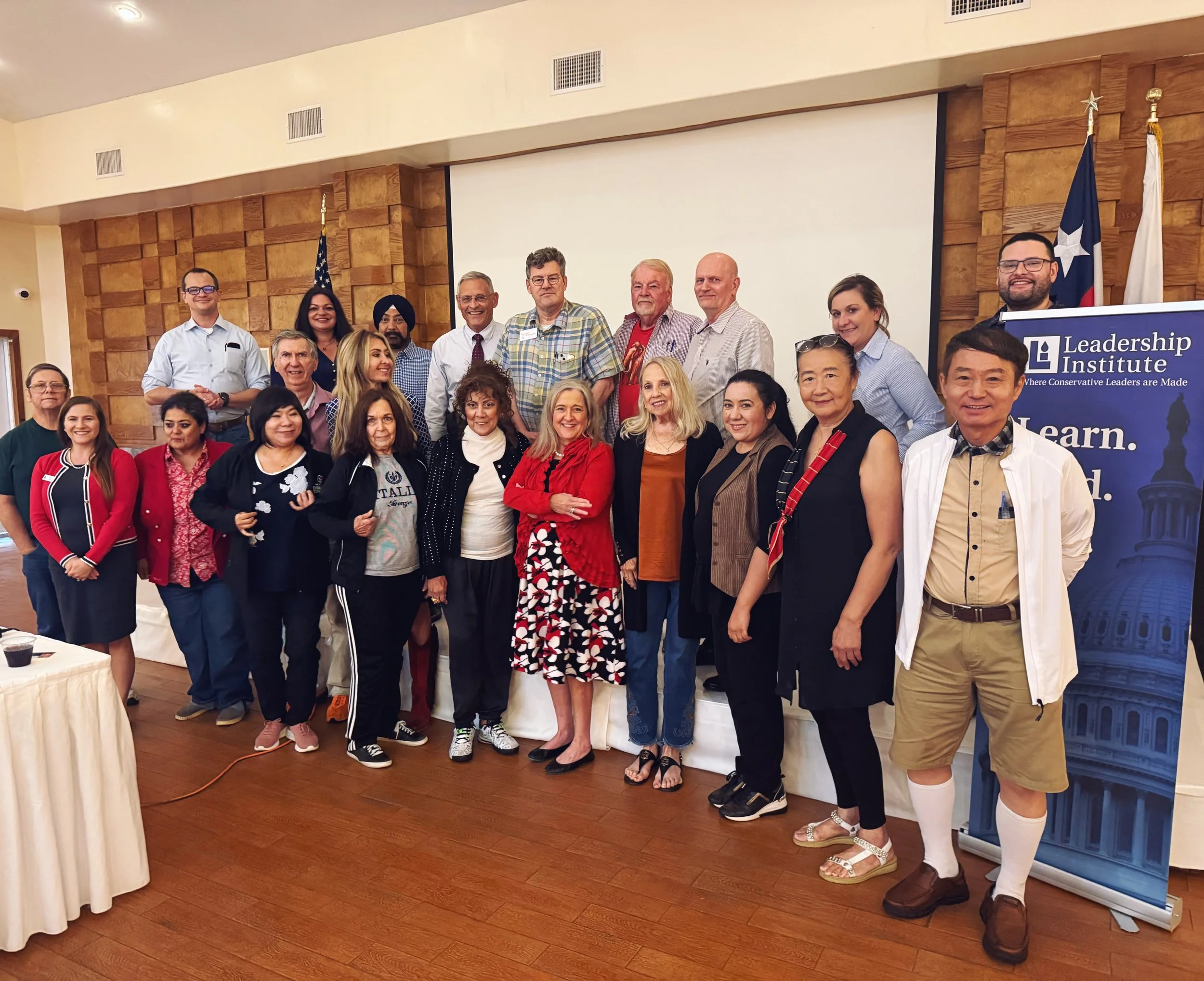Group of diverse adults gathered together in a conference room, posing for a photo during a leadership event, with banners and flags in the background.